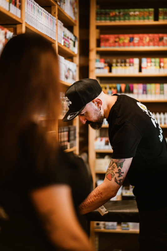 Un uomo con cappello e camicia neri assiste un cliente in un negozio. Sullo sfondo si vedono scaffali pieni di prodotti.