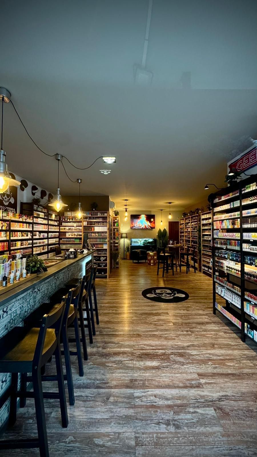 Interno di un negozio, scaffali pieni di prodotti, bar con sgabelli e zona salotto. Pavimenti in legno e luci calde.