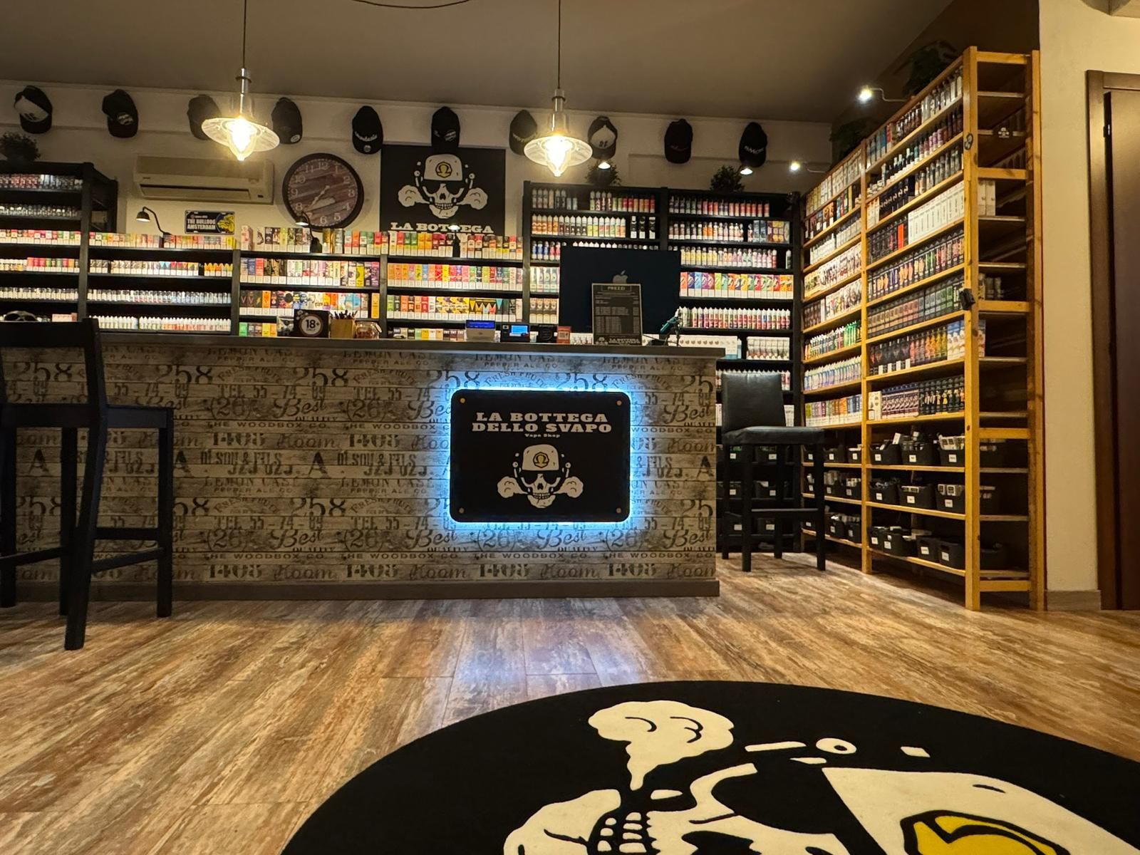 Interno di un negozio di sigarette elettroniche con bancone, scaffali pieni di prodotti e logo con un teschio.
