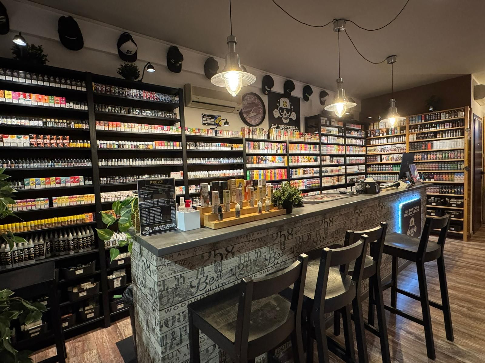 Interno di un negozio di sigarette elettroniche con scaffali pieni di liquidi elettronici, un bar con sgabelli e illuminazione dall'alto.