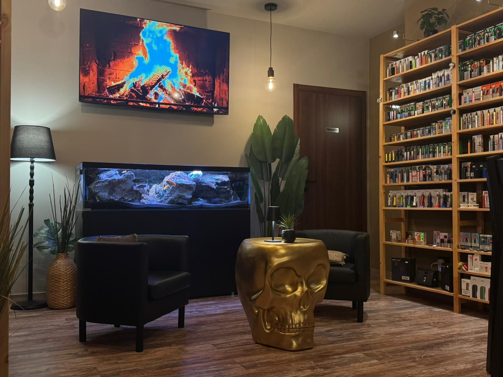 Accogliente area lounge con camino, TV, acquario, sedie nere e tavolo con teschio dorato. Librerie e piante sullo sfondo.