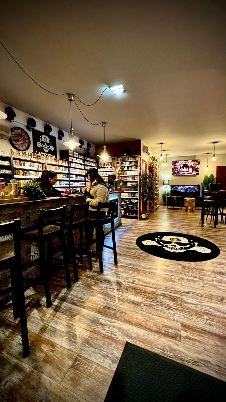 Interno di un negozio di sigarette elettroniche con clienti al bar, scaffali pieni di prodotti e un tappeto con il logo di un teschio sul pavimento.