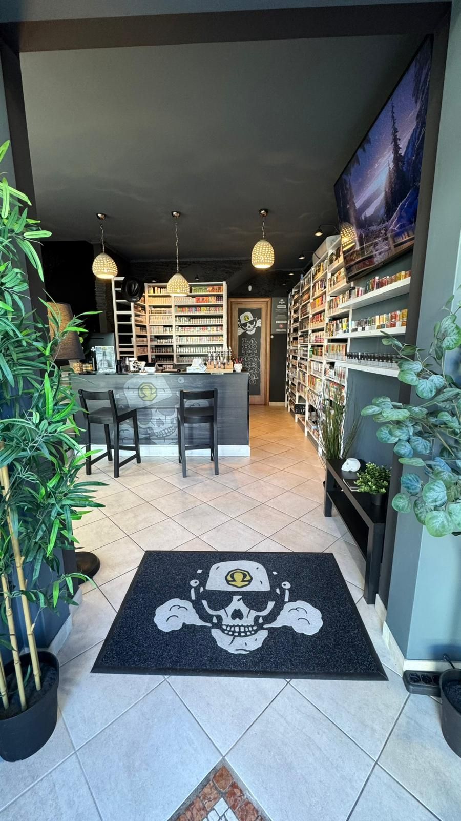 Ingresso di un negozio di sigarette elettroniche, con uno zerbino a forma di teschio, scaffali pieni di prodotti e un bancone con sgabelli.