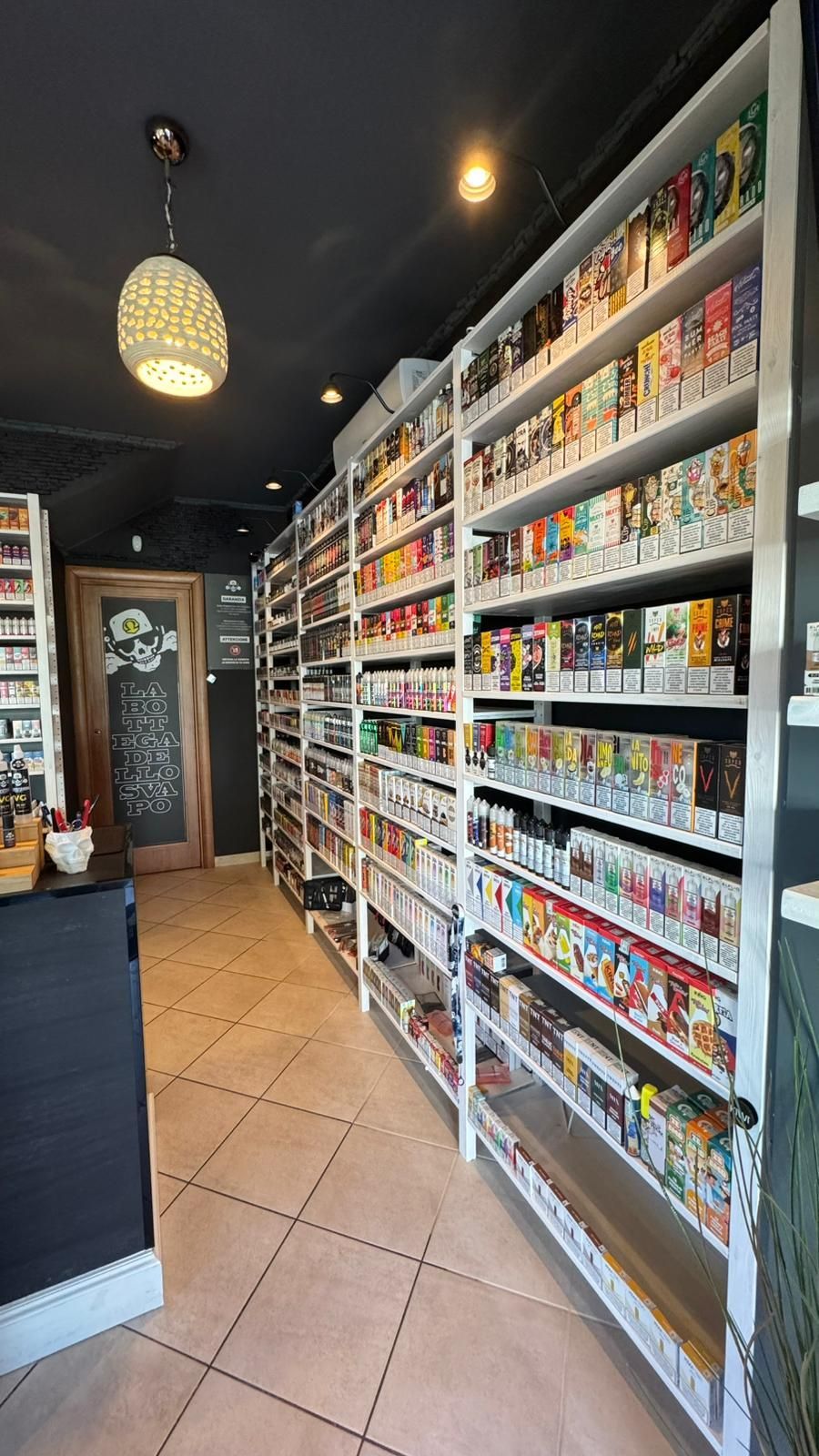 Una corsia di una libreria con alti scaffali bianchi pieni di libri; una lampada è appesa sopra il pavimento piastrellato color marrone chiaro.