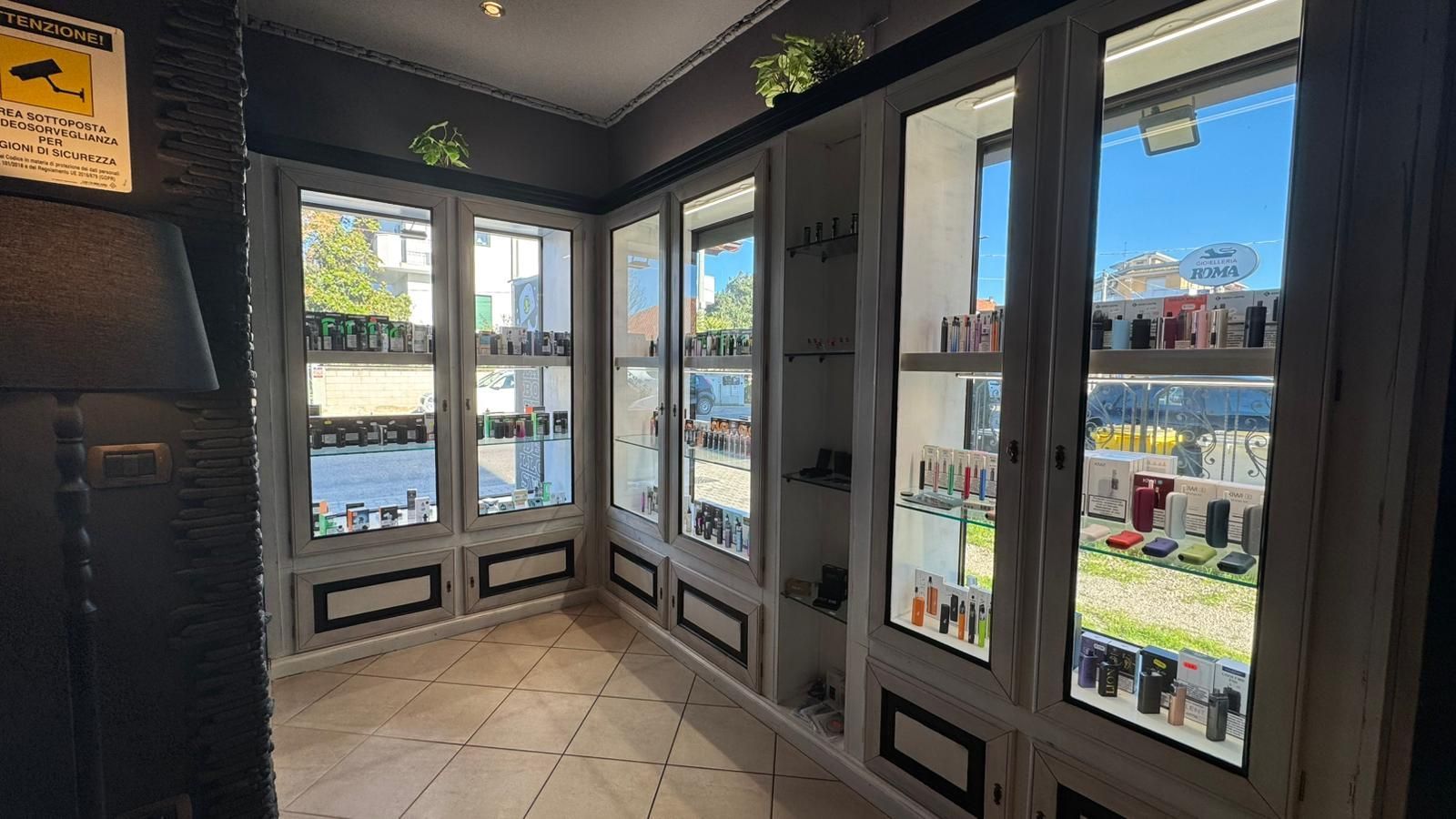 Interno di un negozio con vetrine in vetro piene di prodotti, che si affacciano su una strada soleggiata.