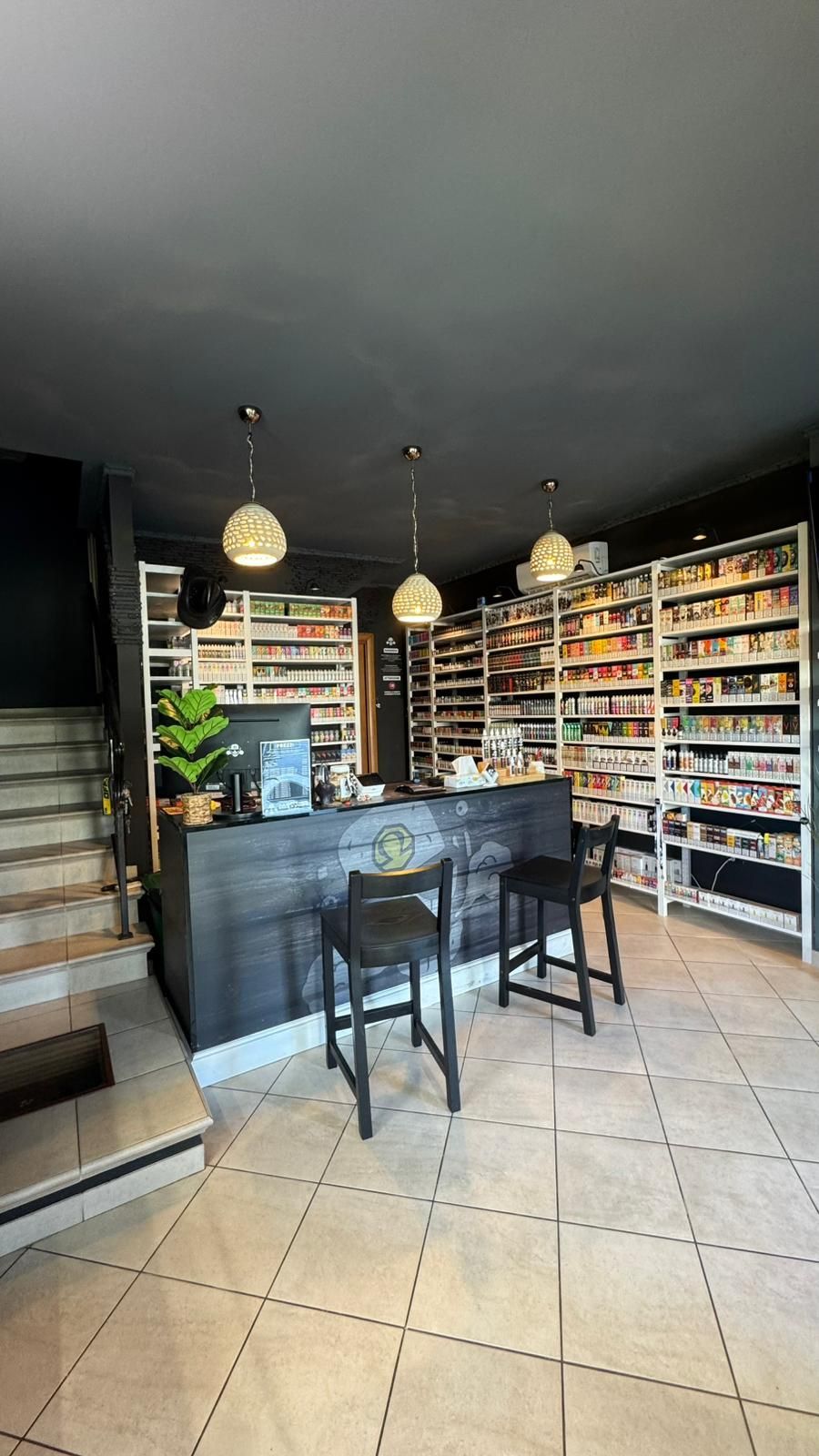 L'interno di un negozio di sigarette elettroniche con un bancone in legno scuro, due sgabelli, scaffali con prodotti e tre lampade a sospensione.