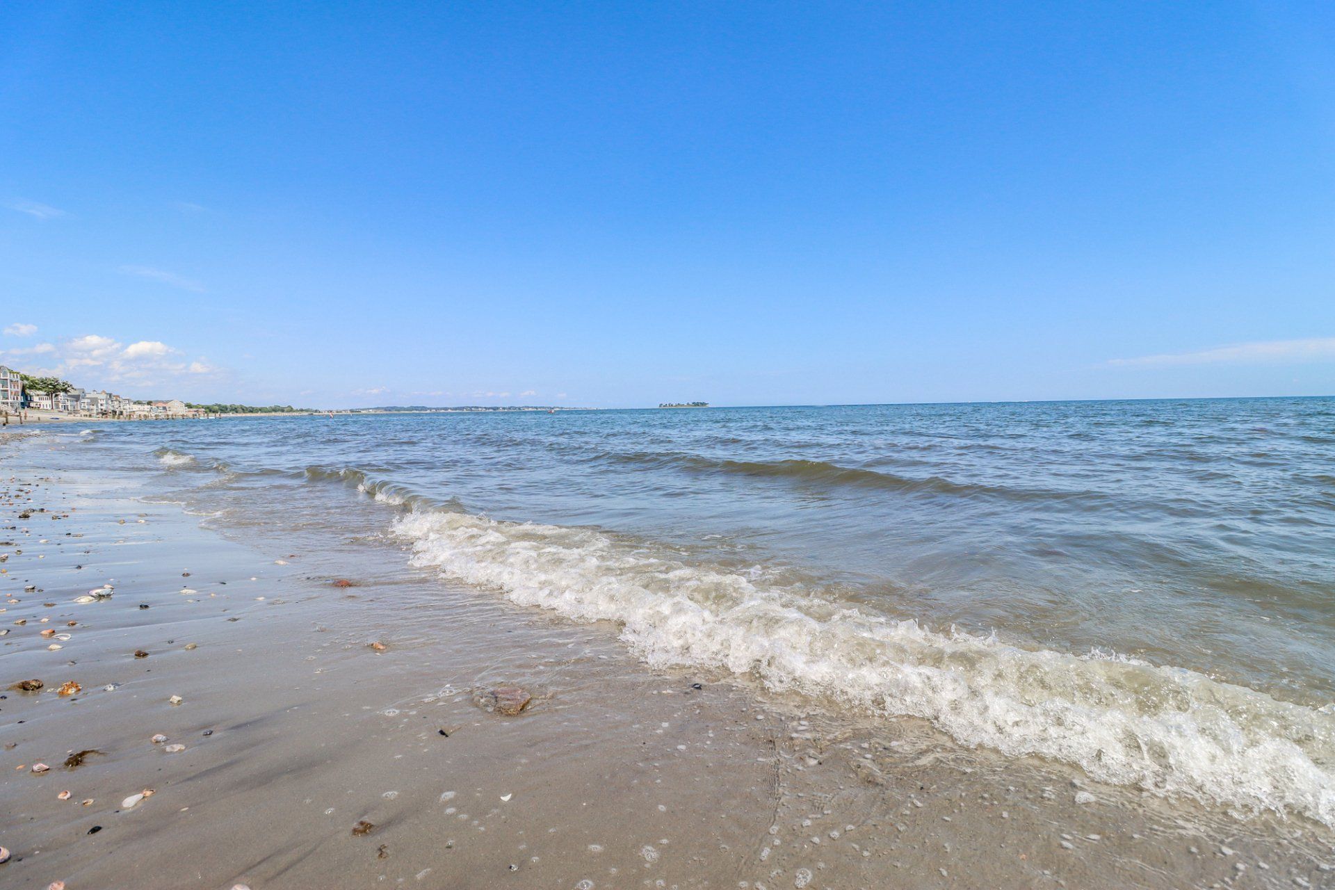 Beach with gentle waves under a clear blue sky, buildings in the distance.