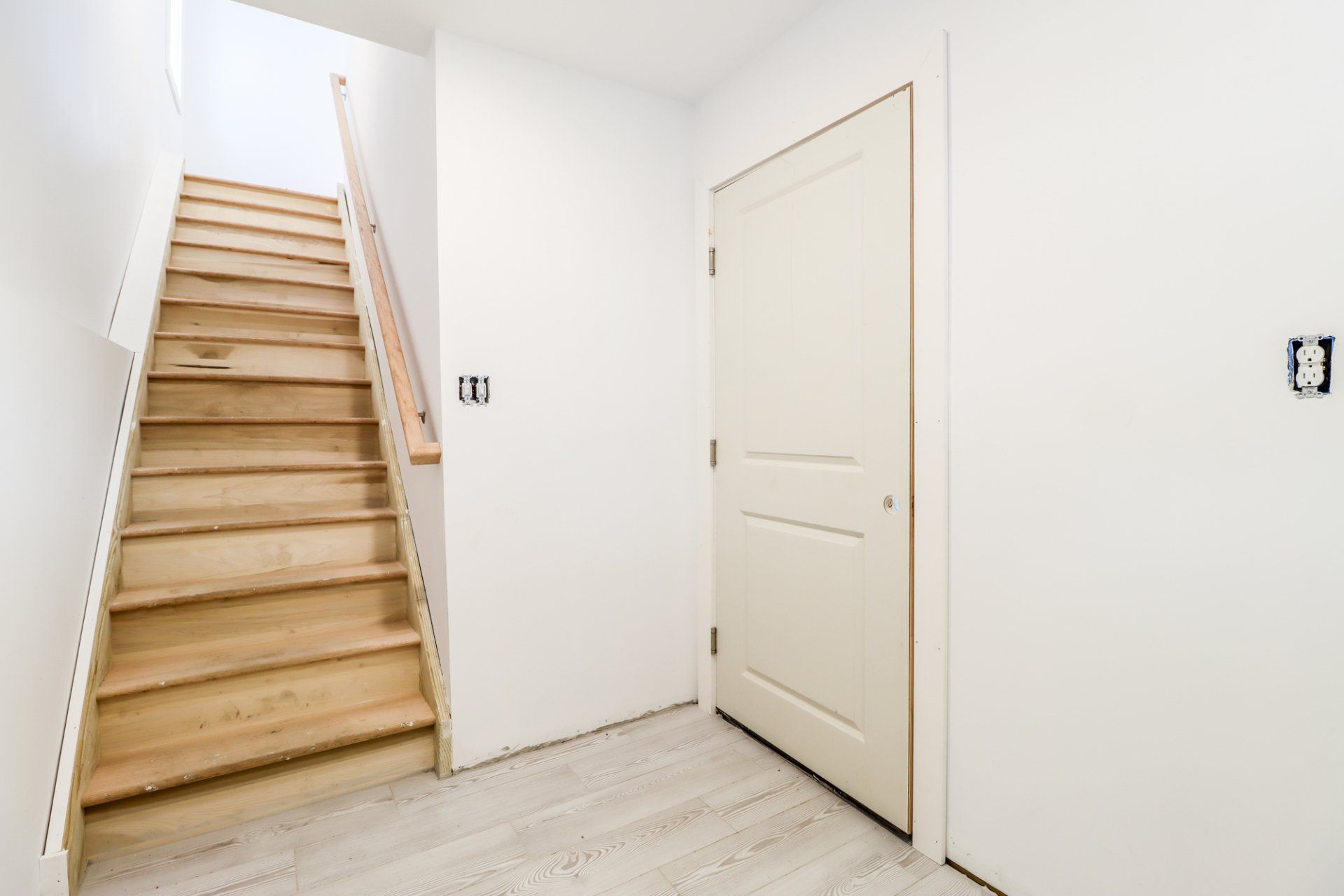 Wooden staircase leading upwards, next to a white door in a white-walled hallway.