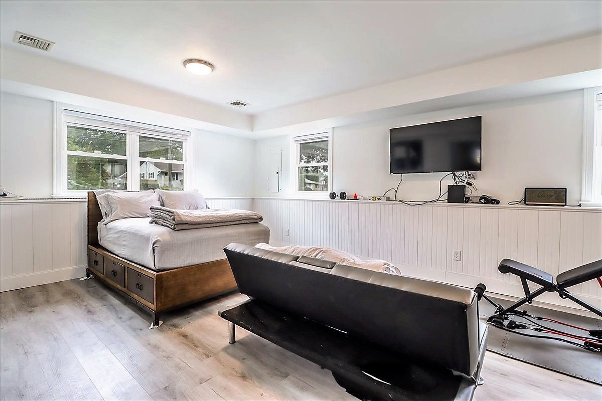 Bedroom with bed, sofa, TV, and exercise equipment. White walls and windows.