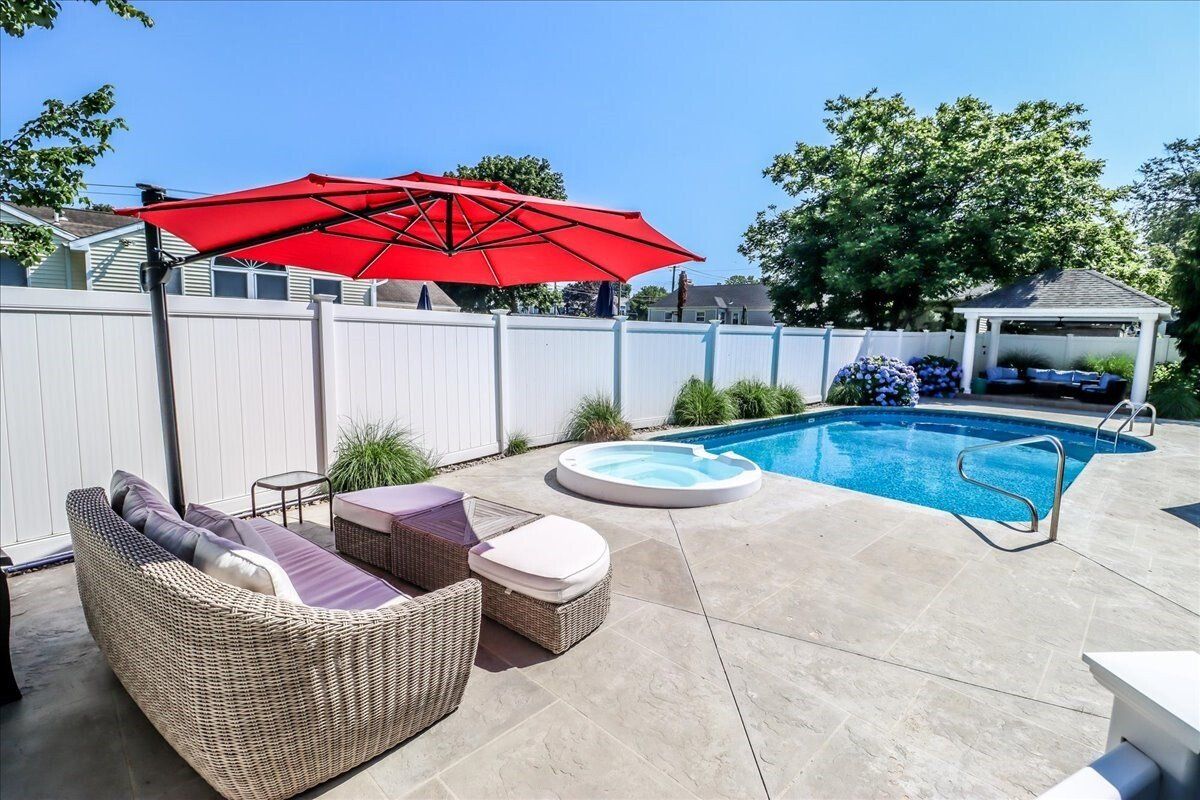 Backyard with a pool, hot tub, and seating area under a red umbrella, enclosed by a white fence.
