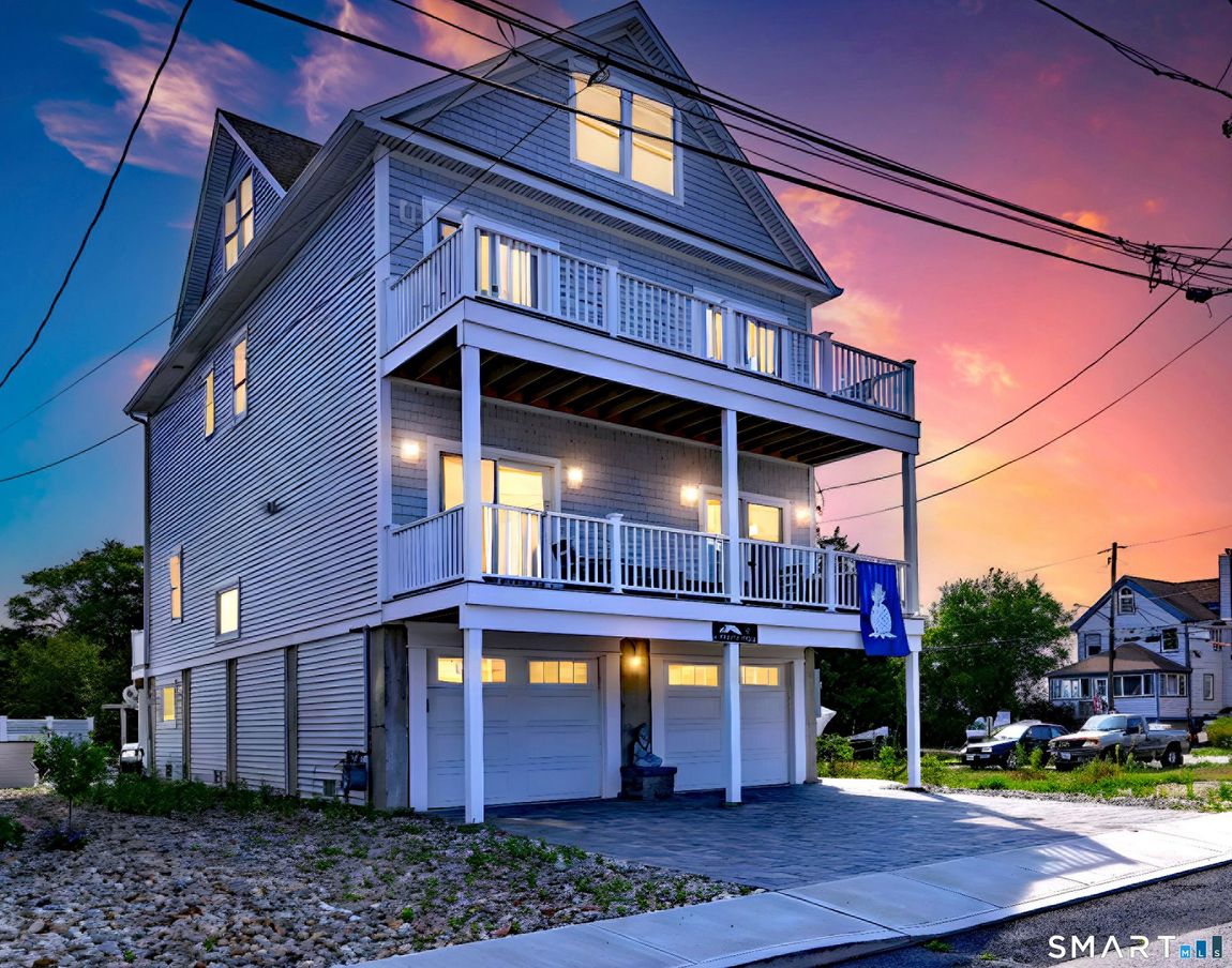 Three-story coastal home with balconies, white siding, and garage doors. Pink and orange sunset sky.