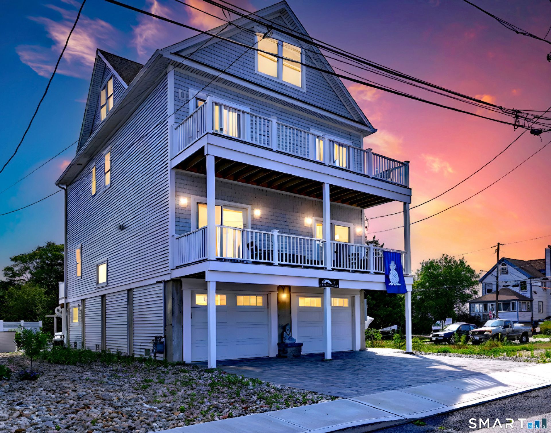 Three-story coastal home with balconies, white siding, and garage doors. Pink and orange sunset sky.