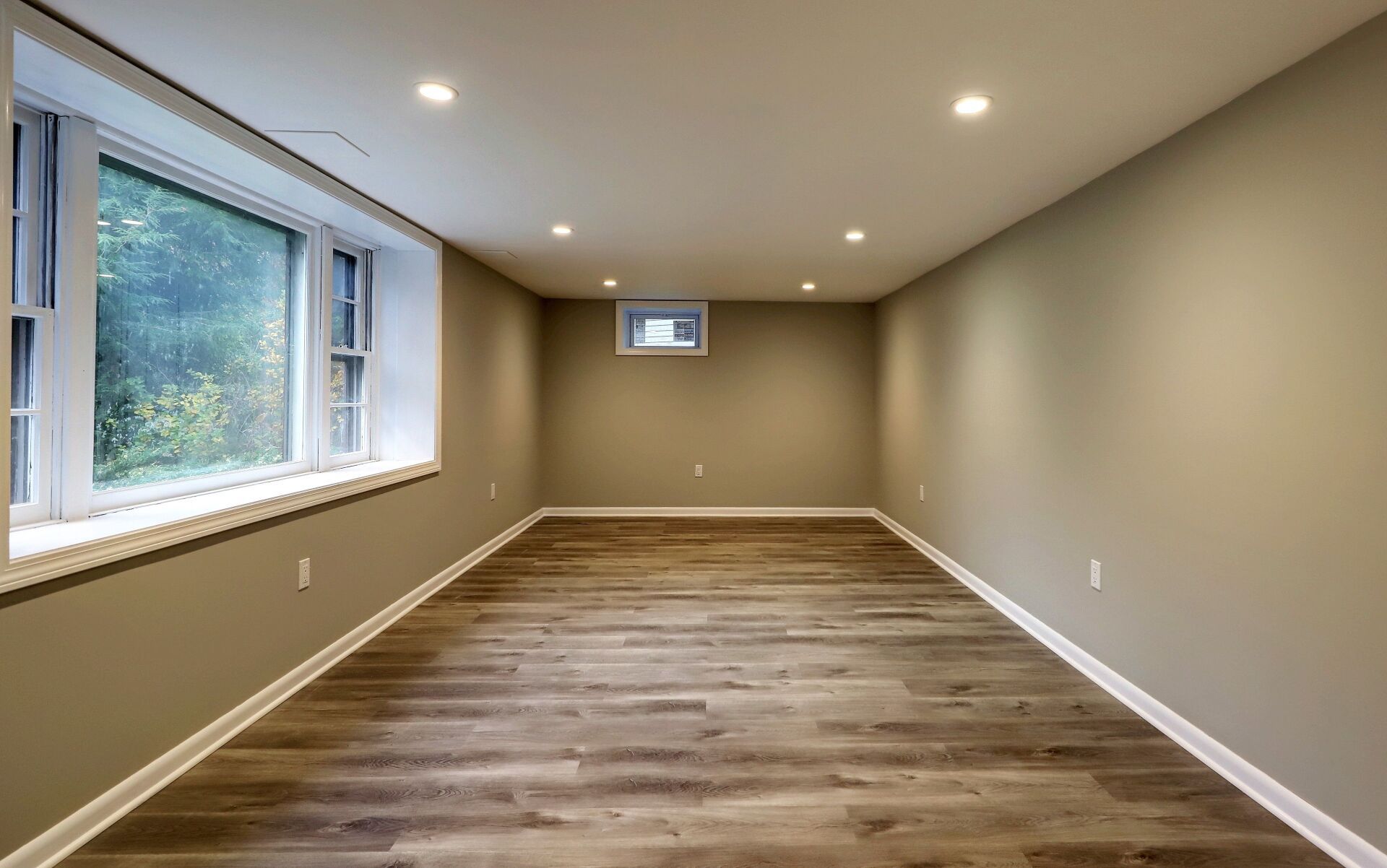 Finished basement with wood-look floor, gray walls, recessed lighting, and a large window with a view of greenery.