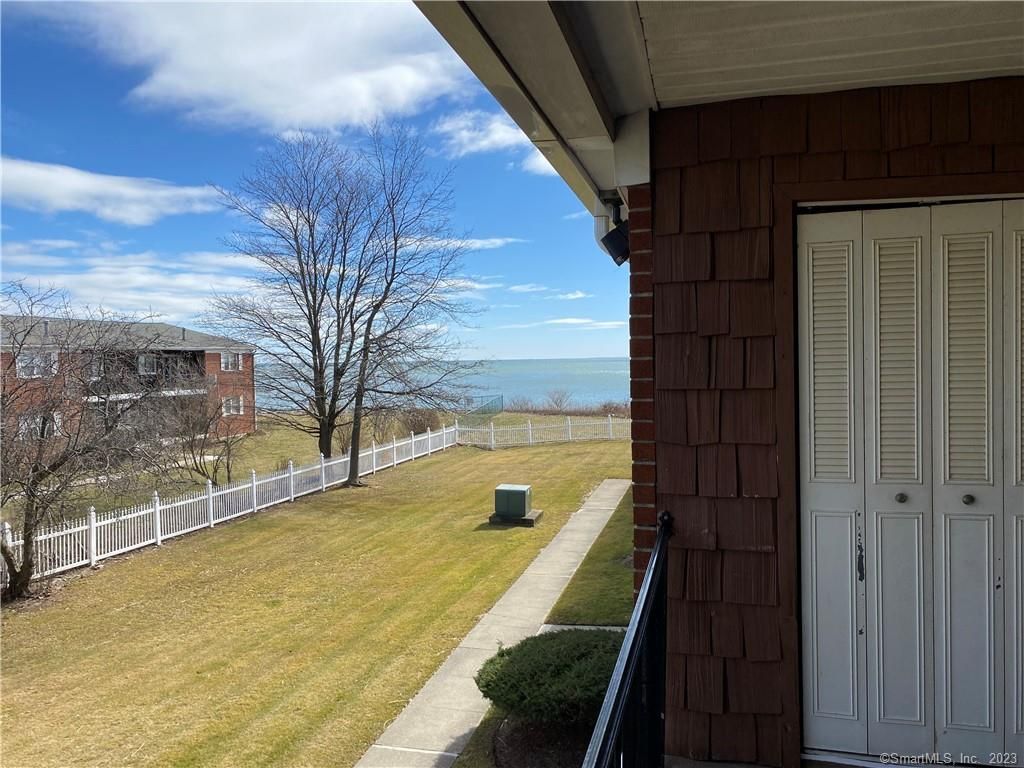 View from a balcony overlooking a grassy yard, white fence, and the ocean under a blue sky.