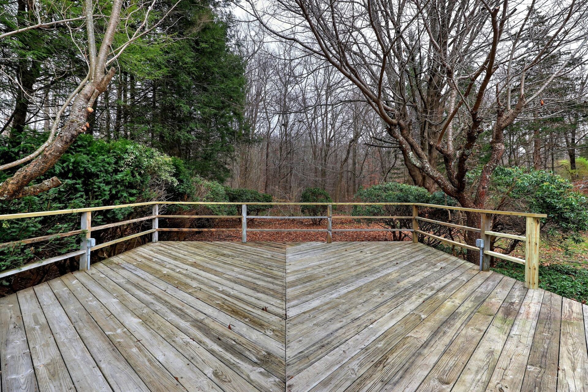 Wooden deck with railing overlooking a wooded area. The deck's wood appears weathered, trees are bare.