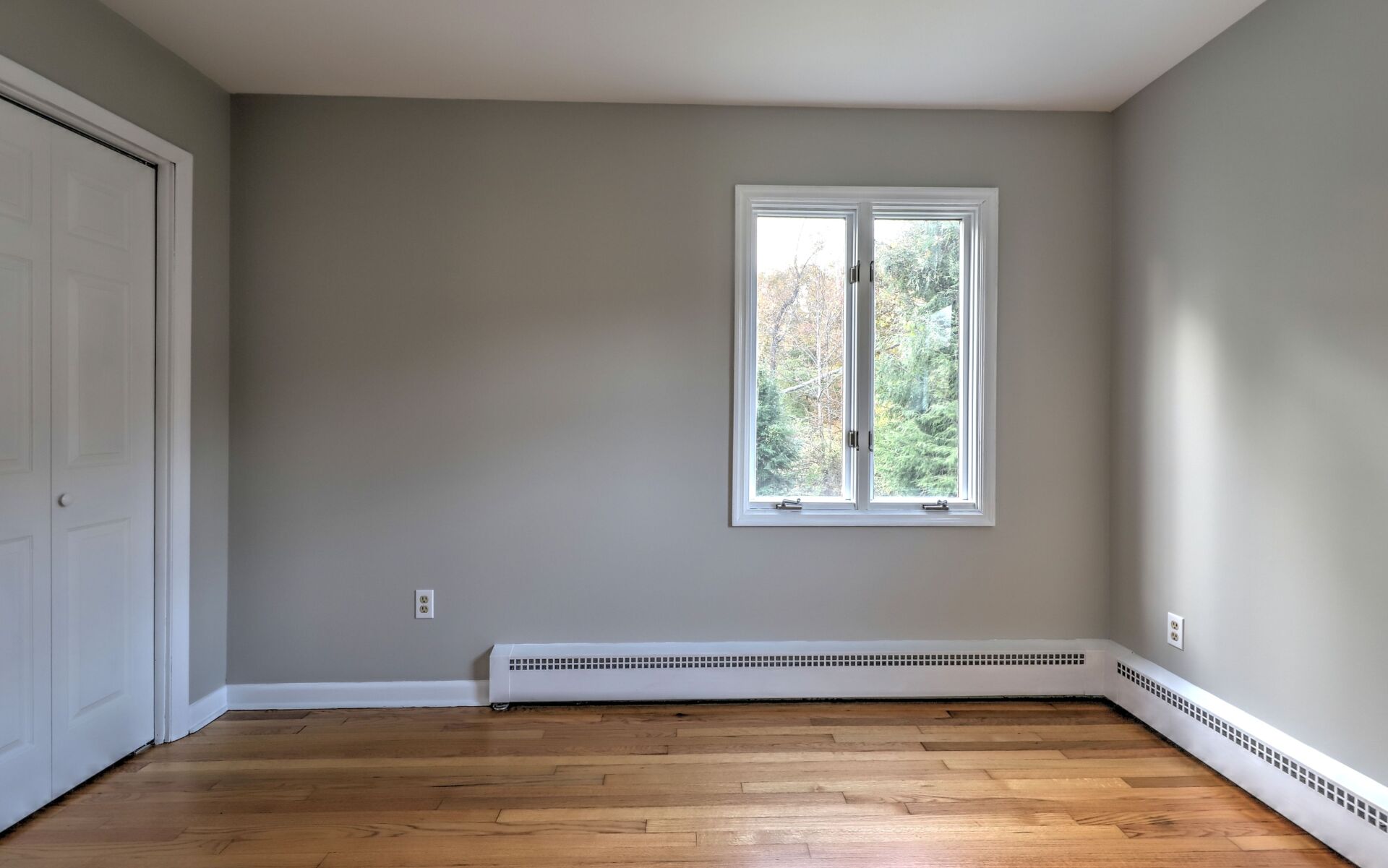 Empty room with hardwood floors, closet, window, and radiator against gray walls.