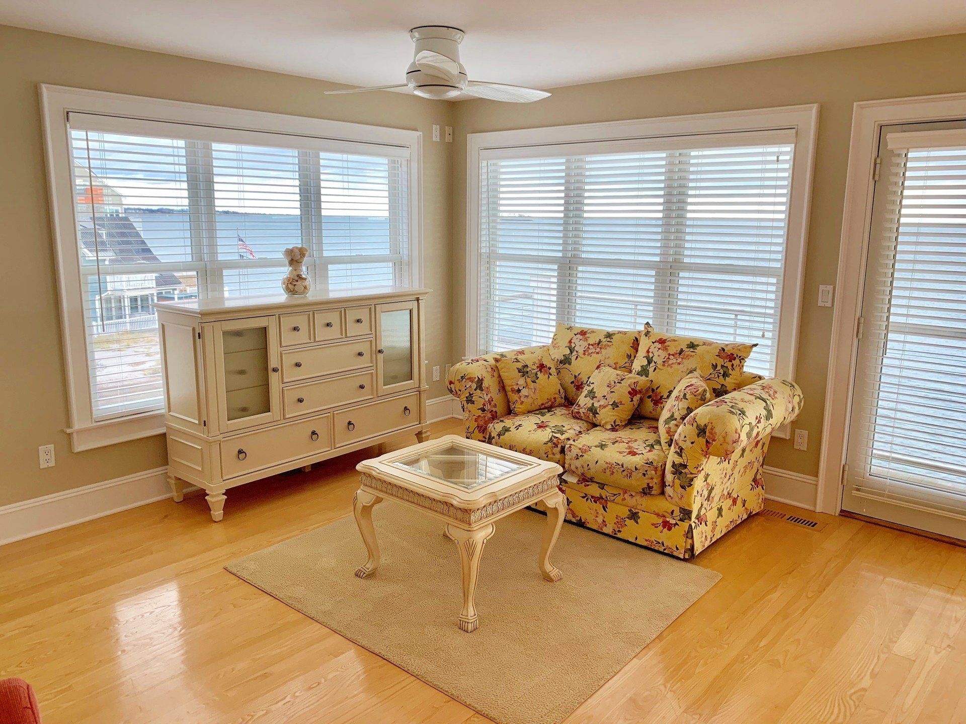Living room with floral sofa, white cabinet, coffee table, and windows overlooking water.