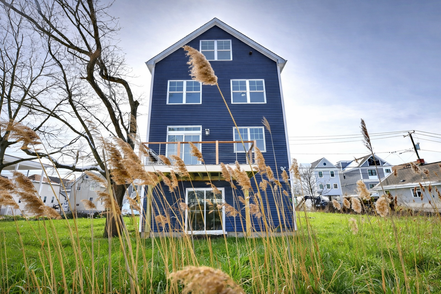 Blue three-story house with deck, in snowy setting, surrounded by tall grass.