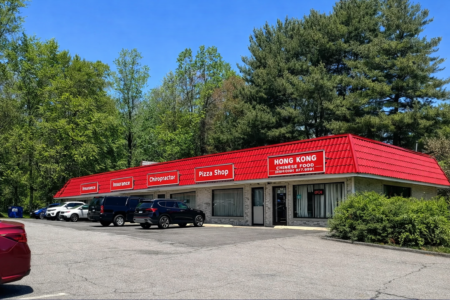 Strip mall with parking area on a sunny day. Trees in the background, blue sky.