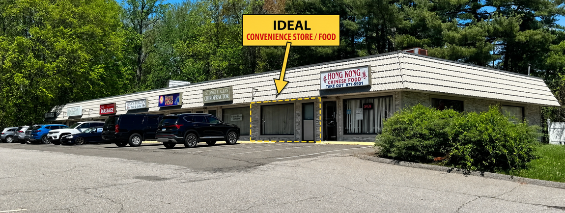 Strip mall with parking area on a sunny day. Trees in the background, blue sky.