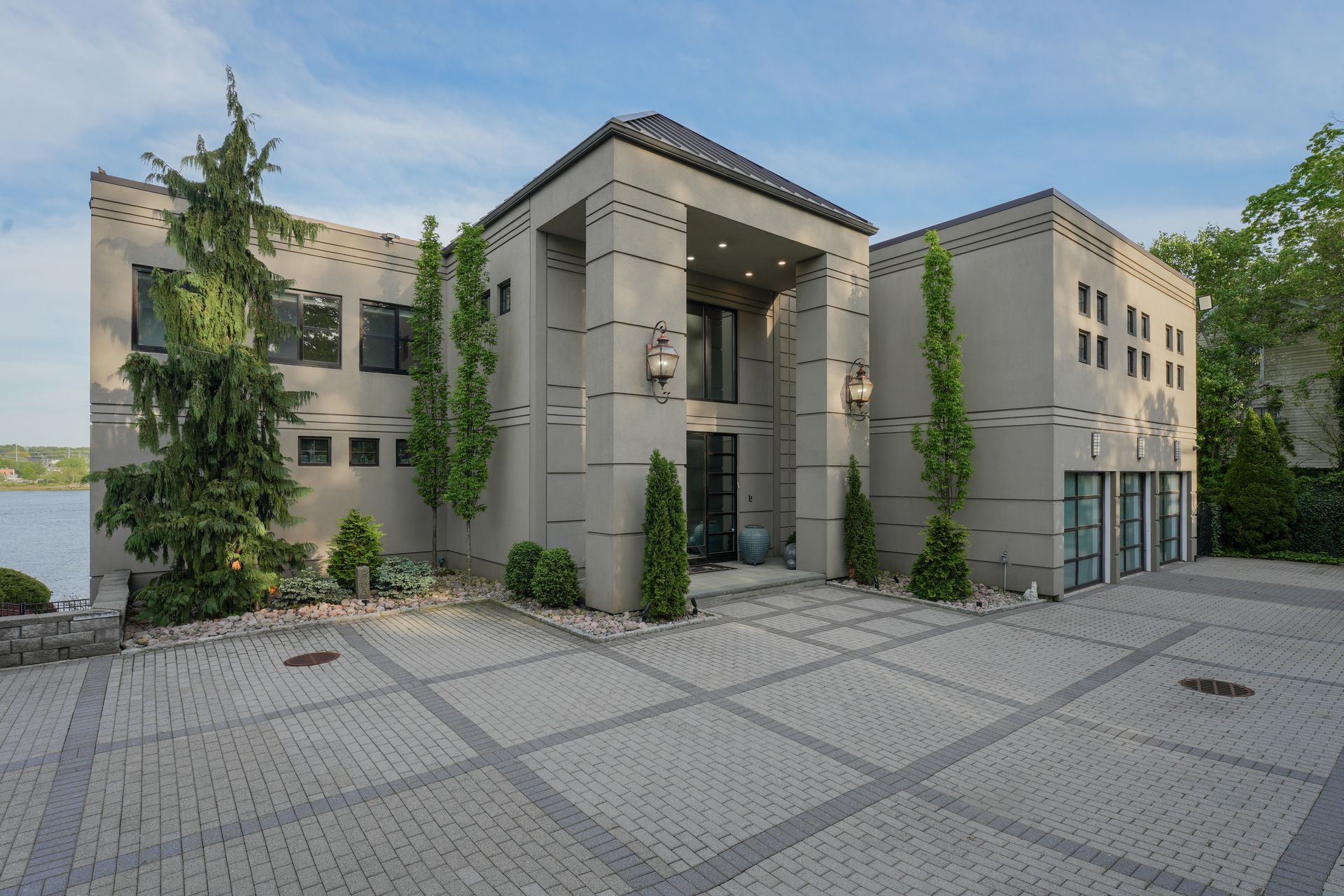 Gray stucco mansion with a paved driveway and trees, facing a body of water.