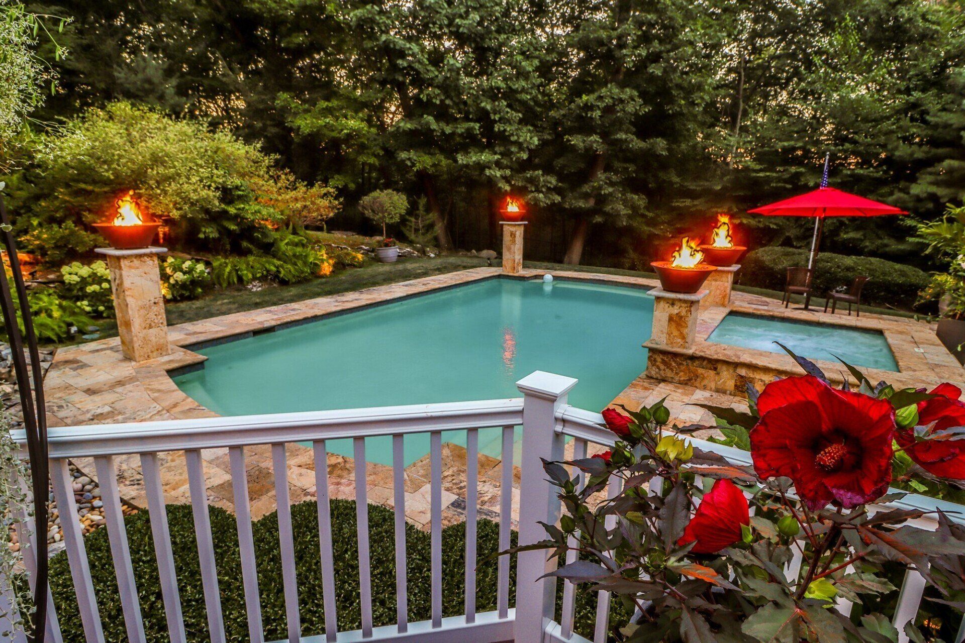 Pool and spa at dusk, with fire bowls, red umbrella, and lush greenery.