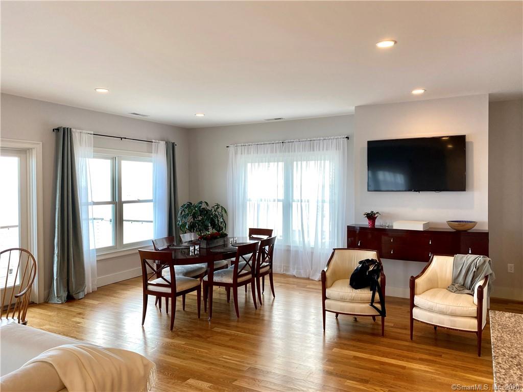 Open-plan living room with dining table, two armchairs, large TV, and windows with sheer curtains. Wooden floors.