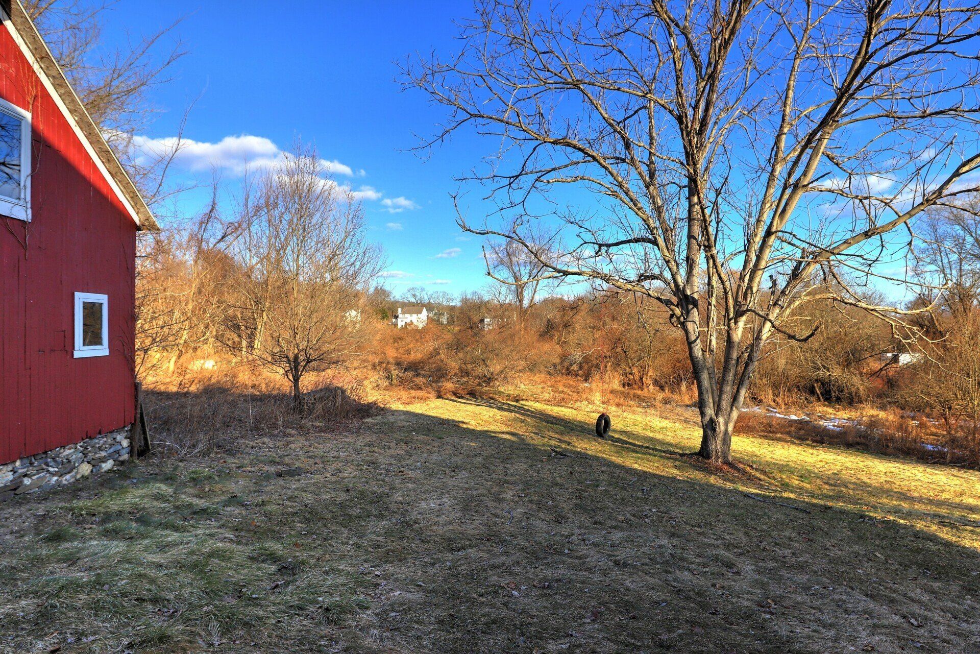 Red barn side with grassy yard, leafless trees under blue sky.