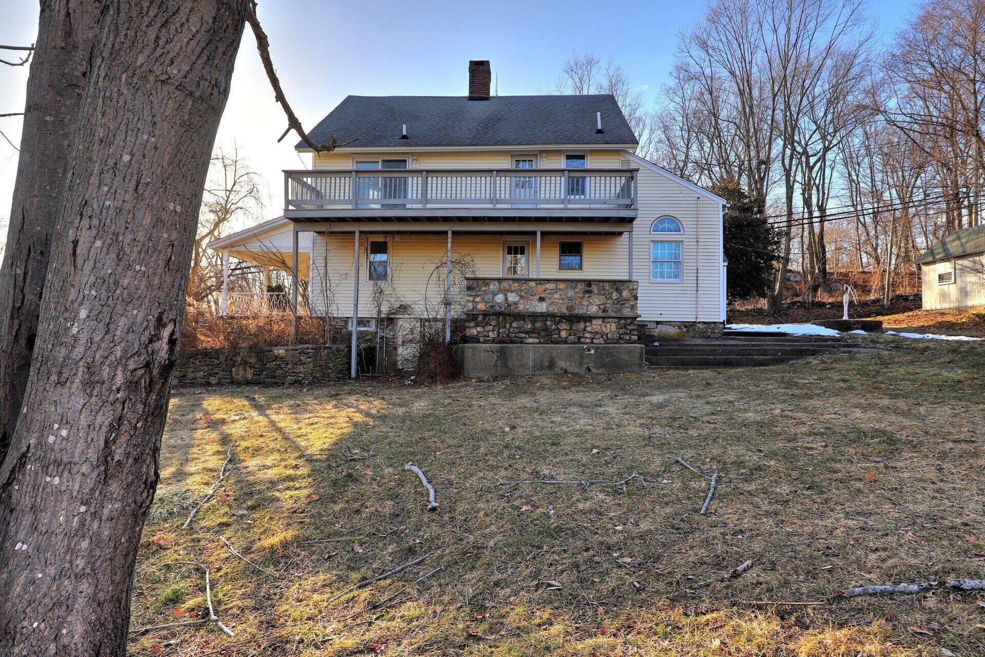 Yellow two-story house with a balcony, stone foundation, and a bare tree in the foreground.