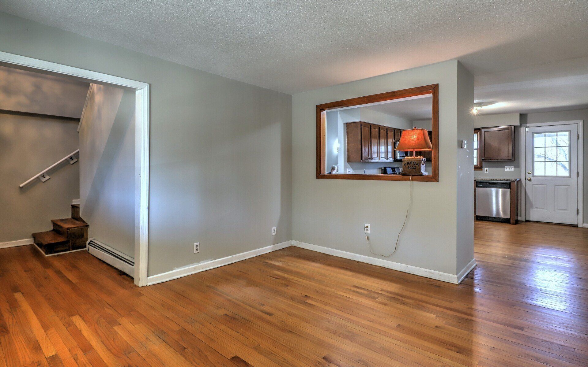 Empty living room with wood floors, doorway to stairs, and a window looking into a kitchen.