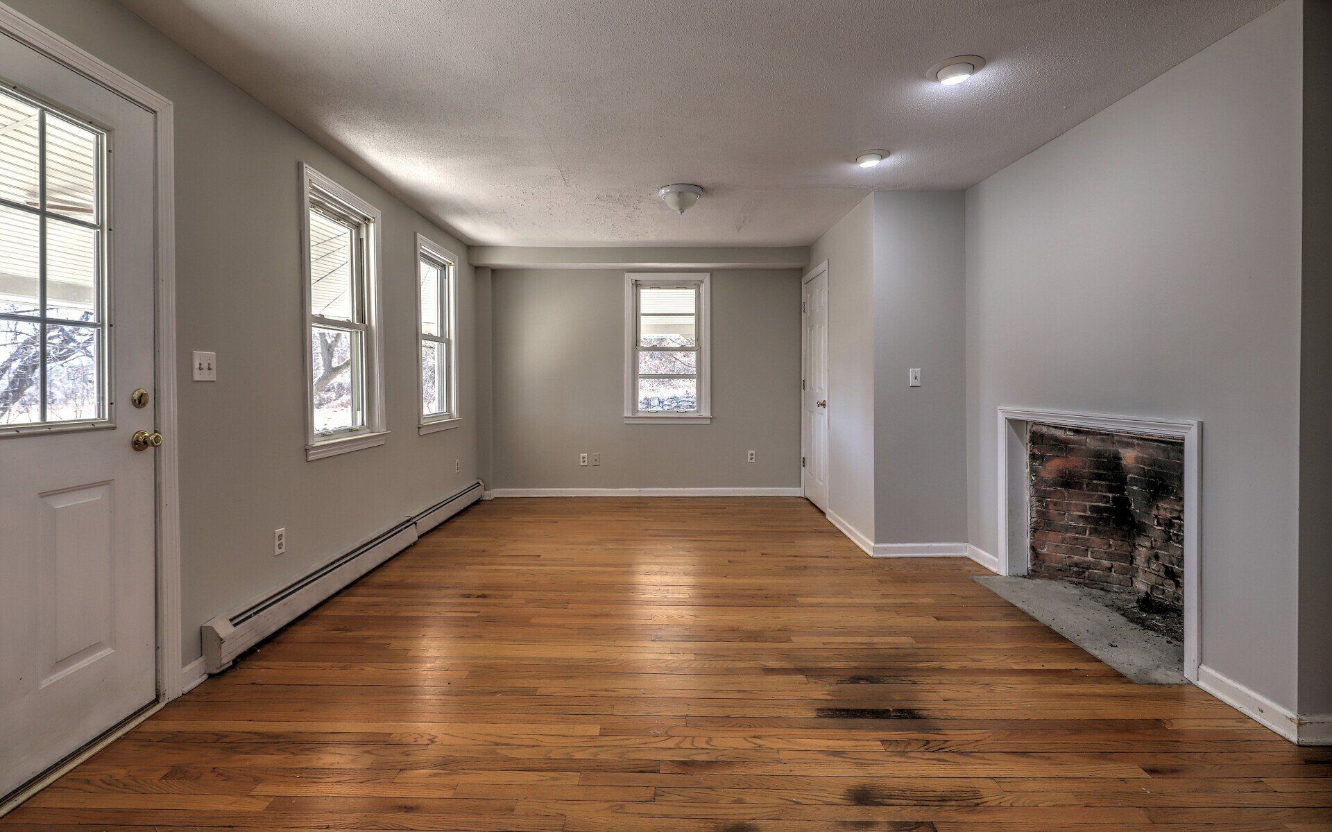 Empty room with hardwood floors, gray walls, windows, fireplace, and door.