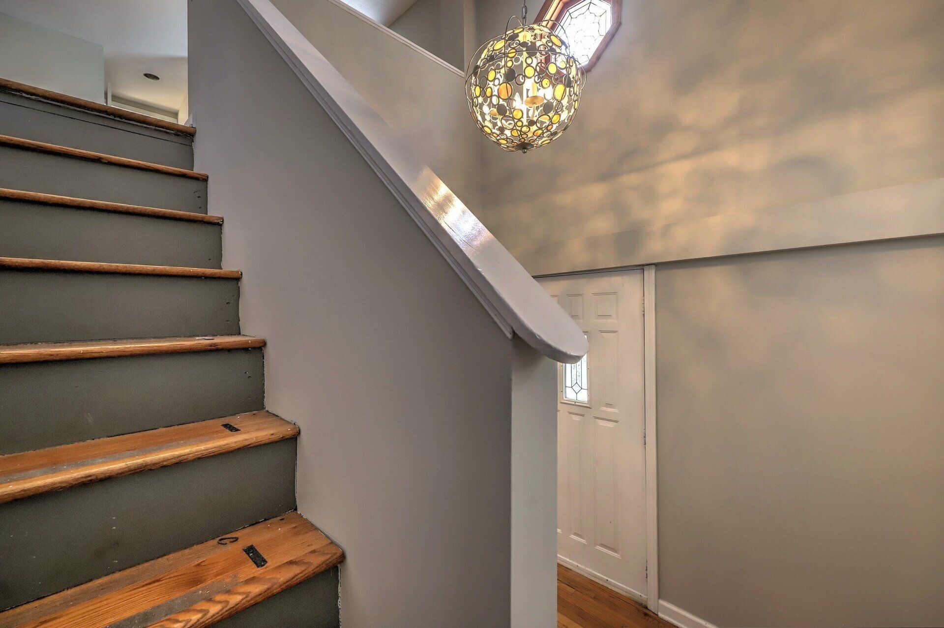 Gray staircase with wooden steps, leading to a white door. A decorative light fixture hangs above.