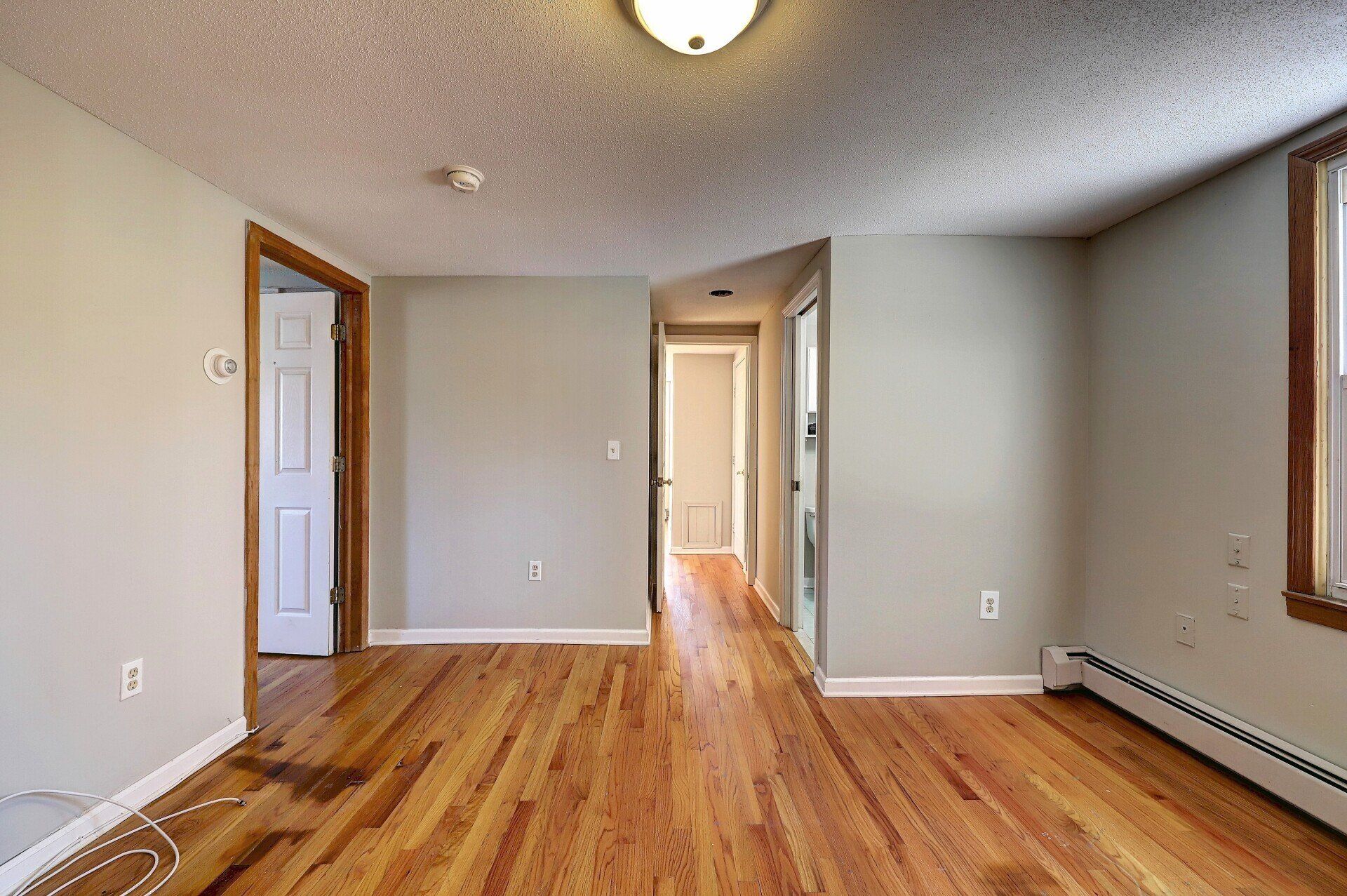 Wooden-floored room with doors, tan walls, and a hallway. White ceiling with a light fixture.