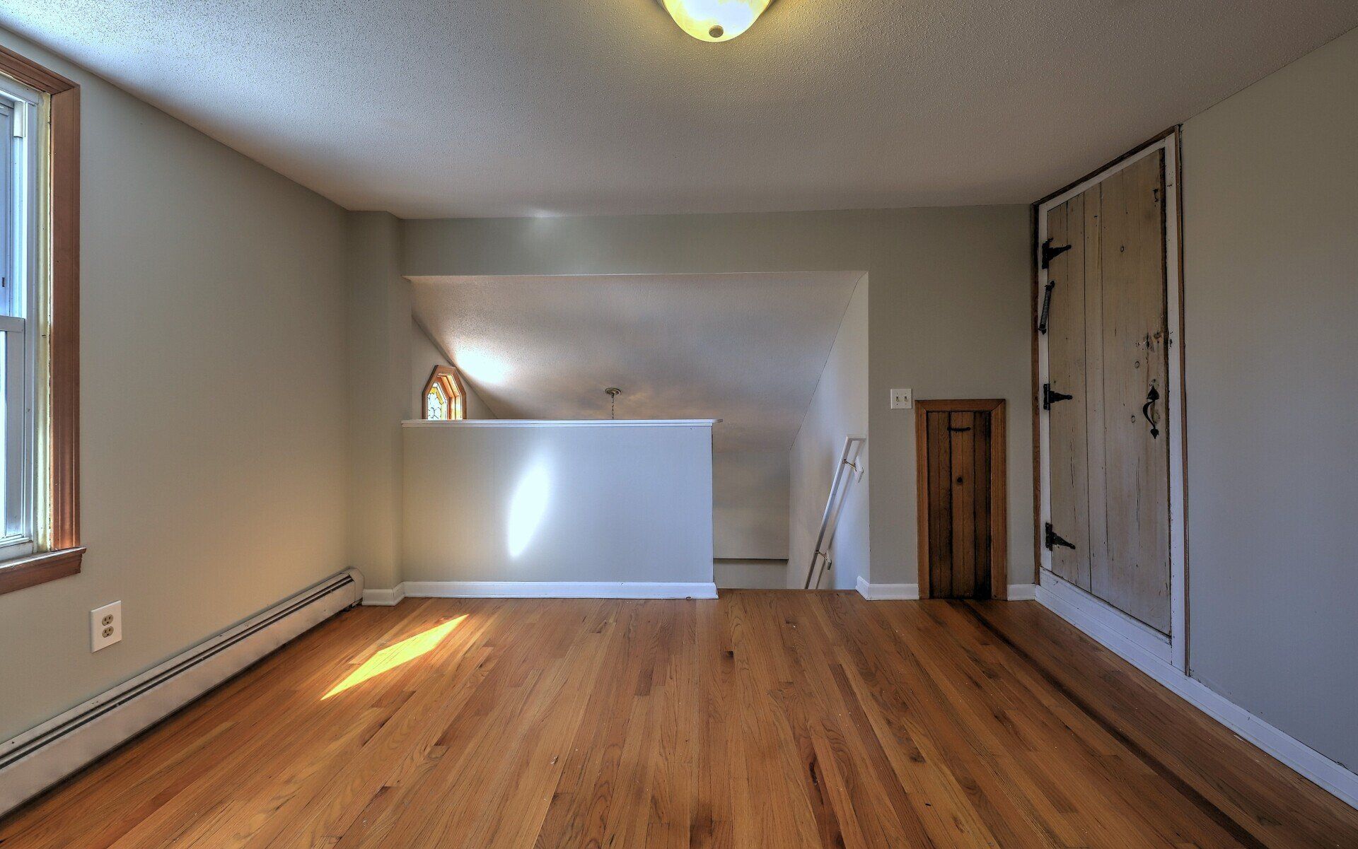 Empty room with hardwood floors, a small window, and a wooden door. Sunlight streams in.