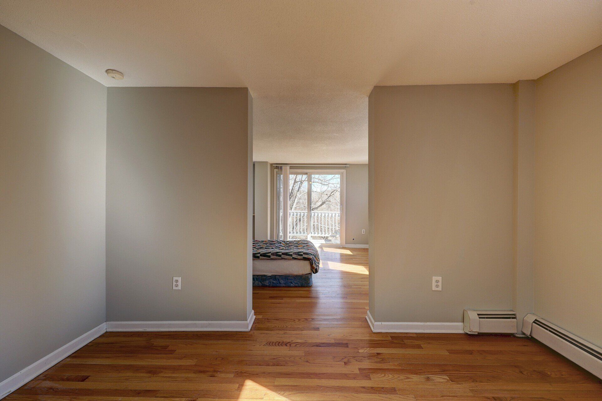 Empty room with hardwood floors, doorways, and a bedroom in the distance.