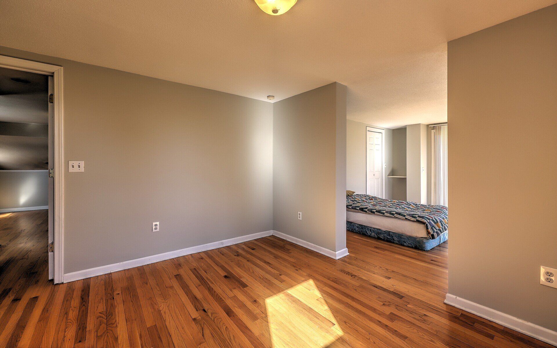 Empty room with hardwood floors, gray walls, and an open doorway to a bedroom.