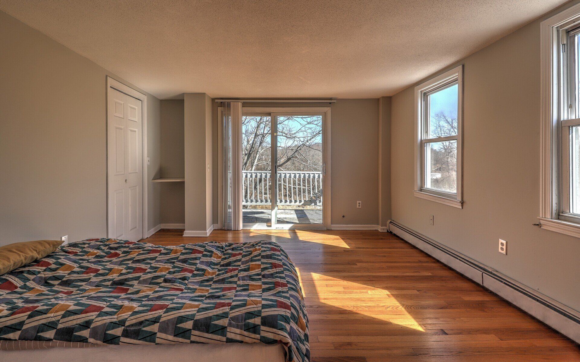 Bedroom with wooden floors, neutral walls, sliding door to a balcony, two windows, and a bed.