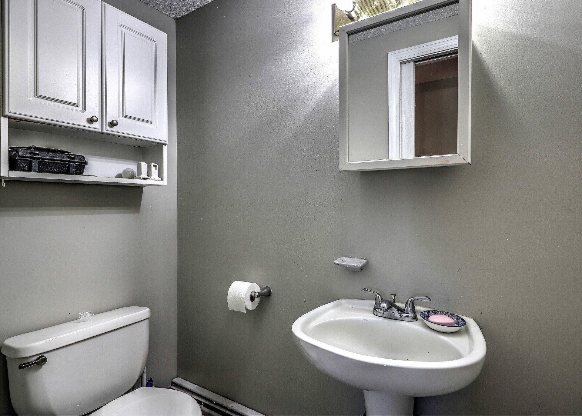 A small bathroom with a toilet, sink, mirror, and cabinet. Gray walls and white fixtures.