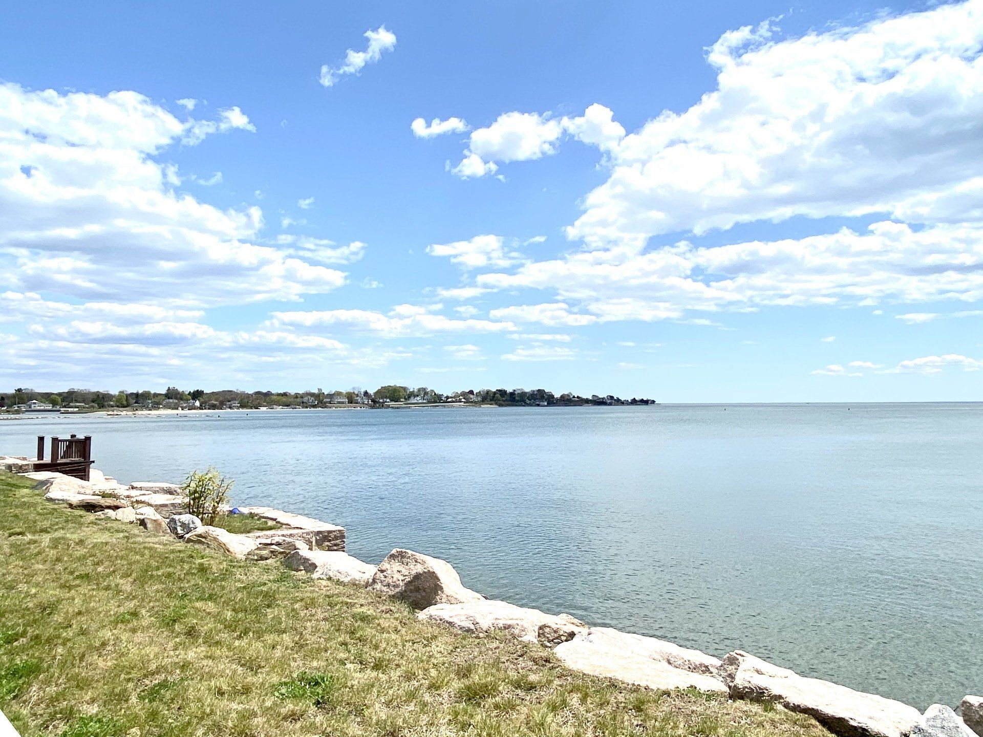 Calm blue lake under a bright sky with puffy clouds. Shoreline with grassy area and rocks.