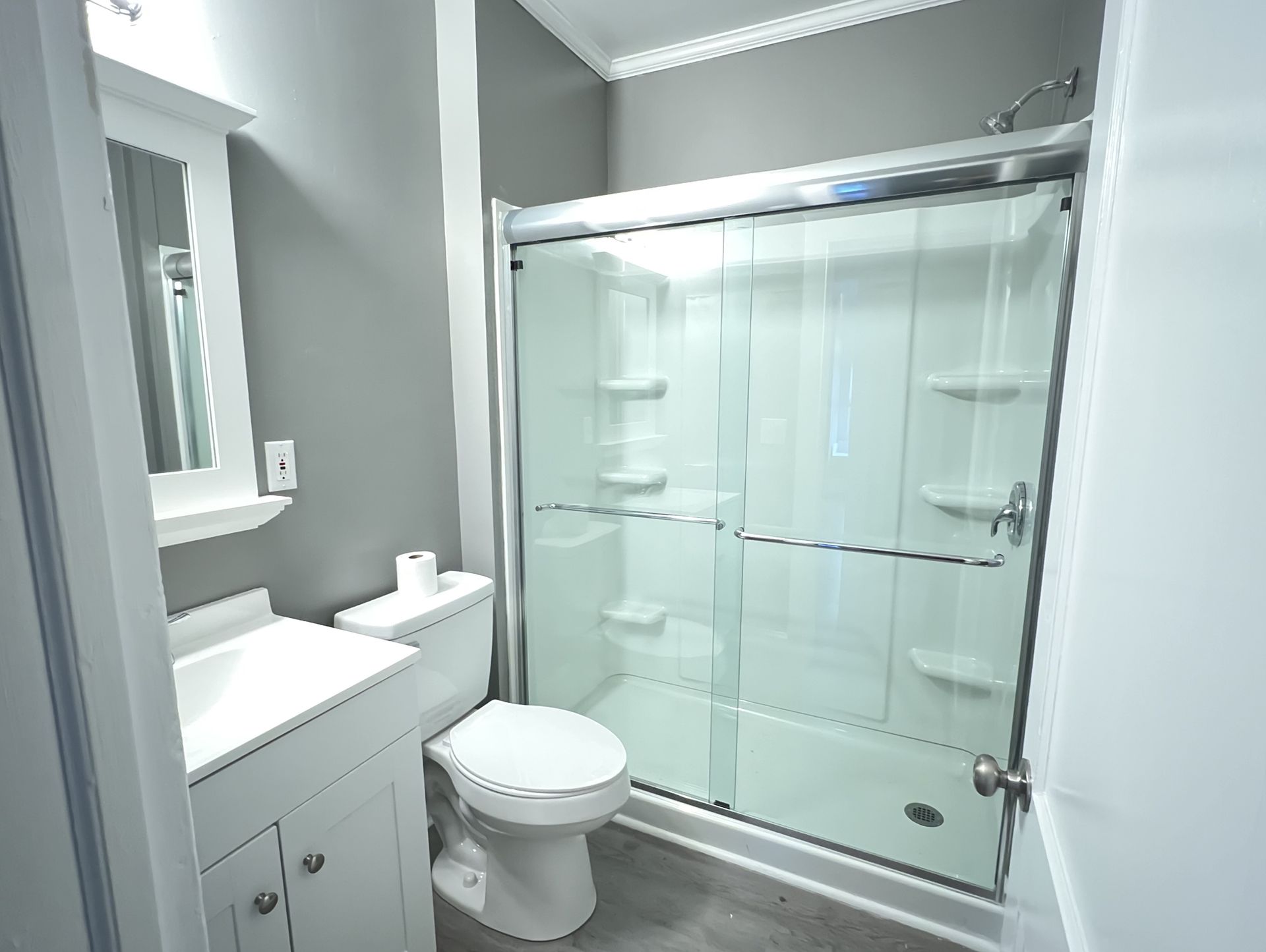 A small, modern bathroom with a shower, toilet, and vanity. Gray walls and white fixtures.