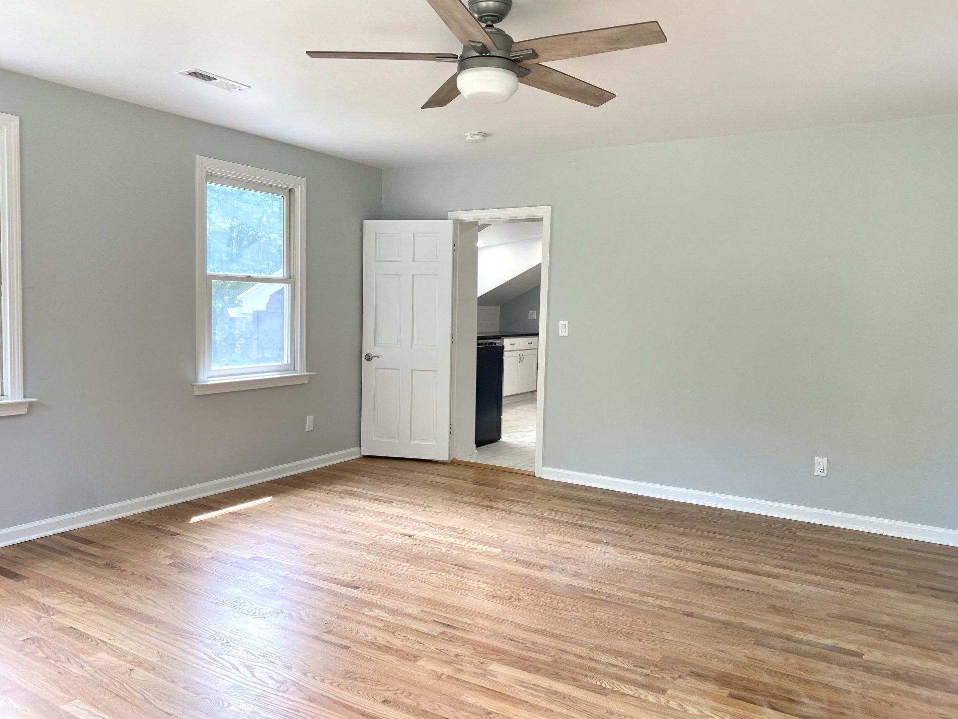 Empty room with hardwood floors, gray walls, open doorway, ceiling fan, and window.