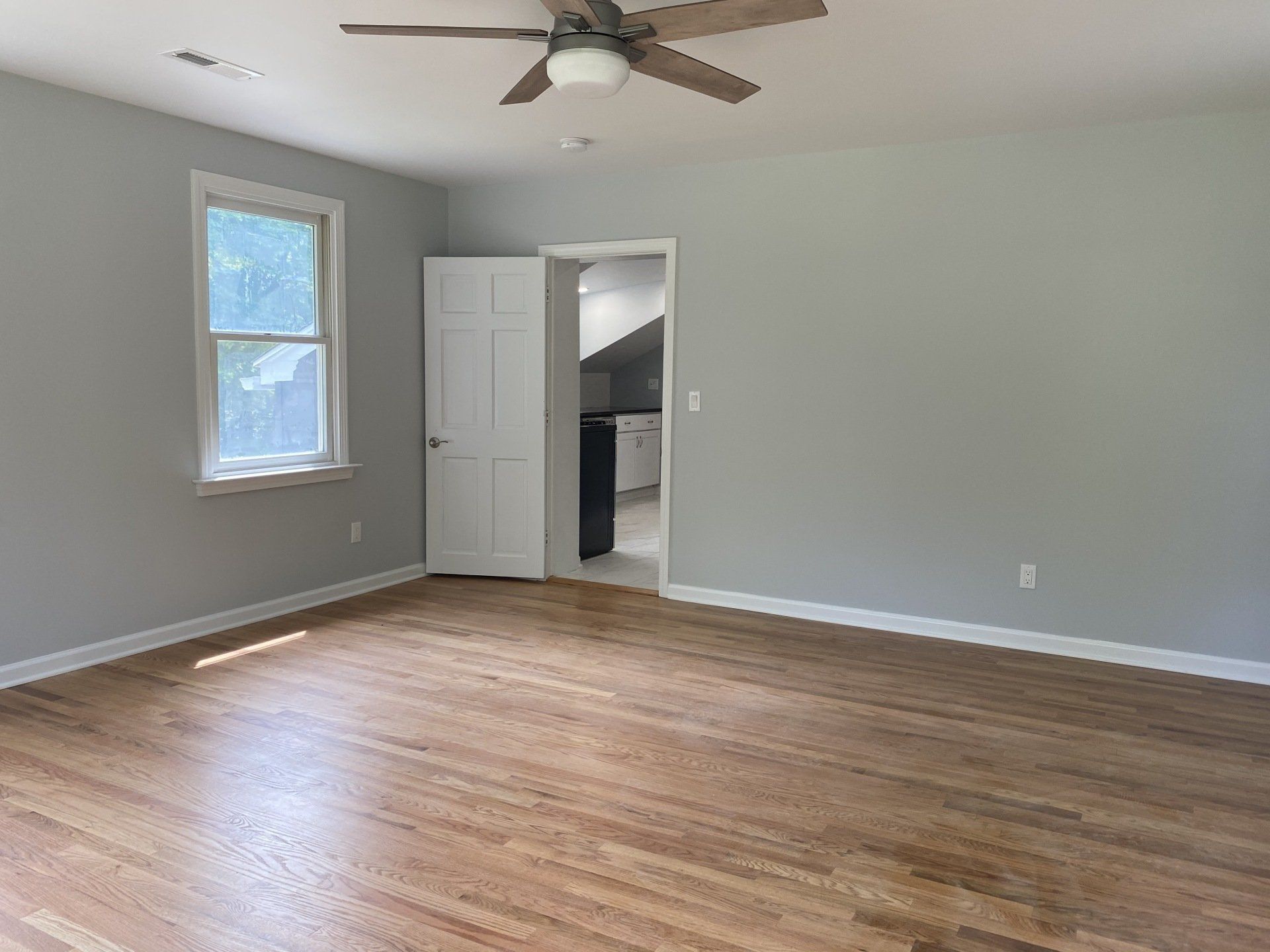 Empty room with light wood floors, grey walls, white door and window, and a ceiling fan.