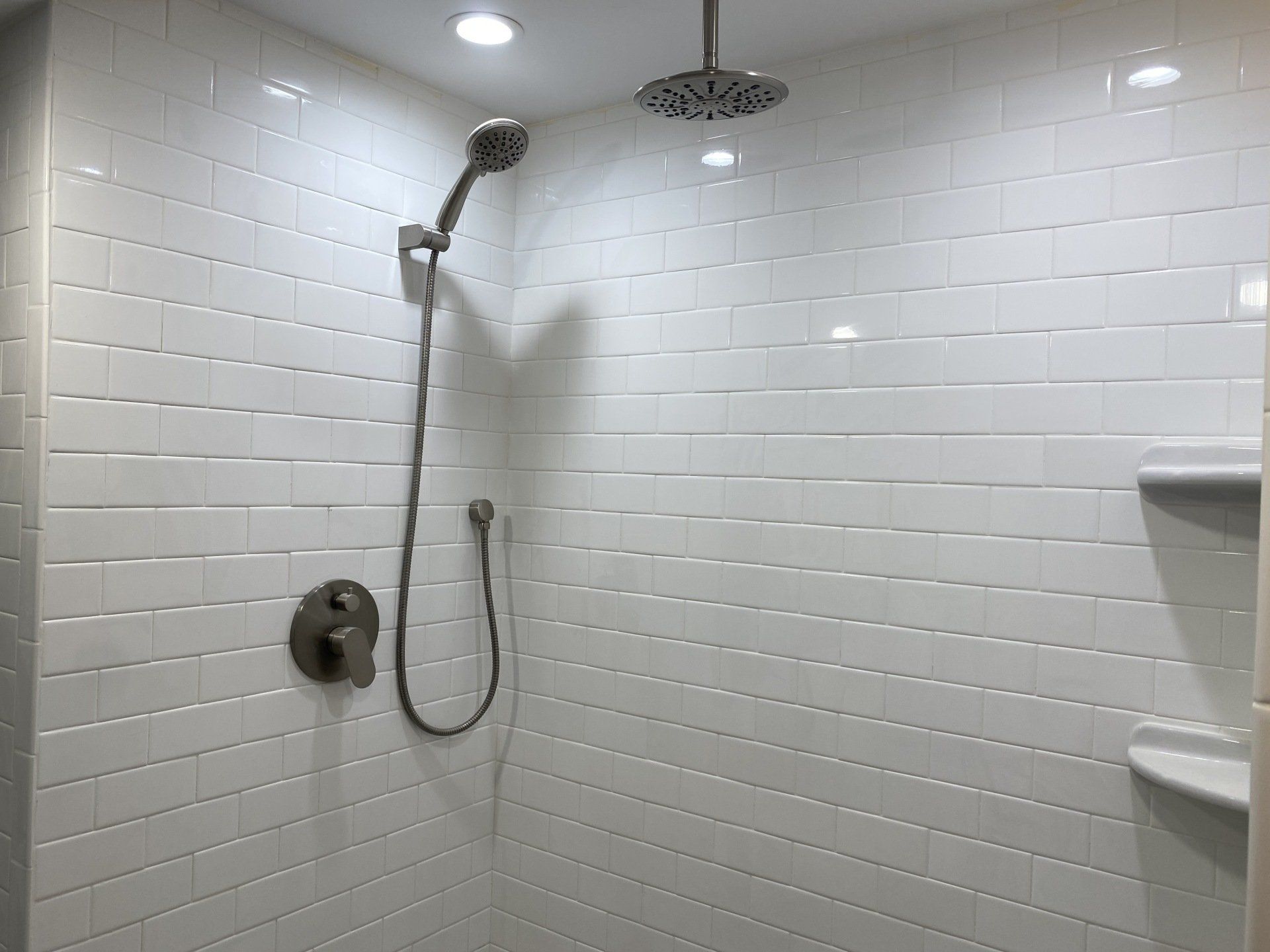White-tiled shower with handheld and overhead showerheads, recessed shelf, and chrome fixtures.