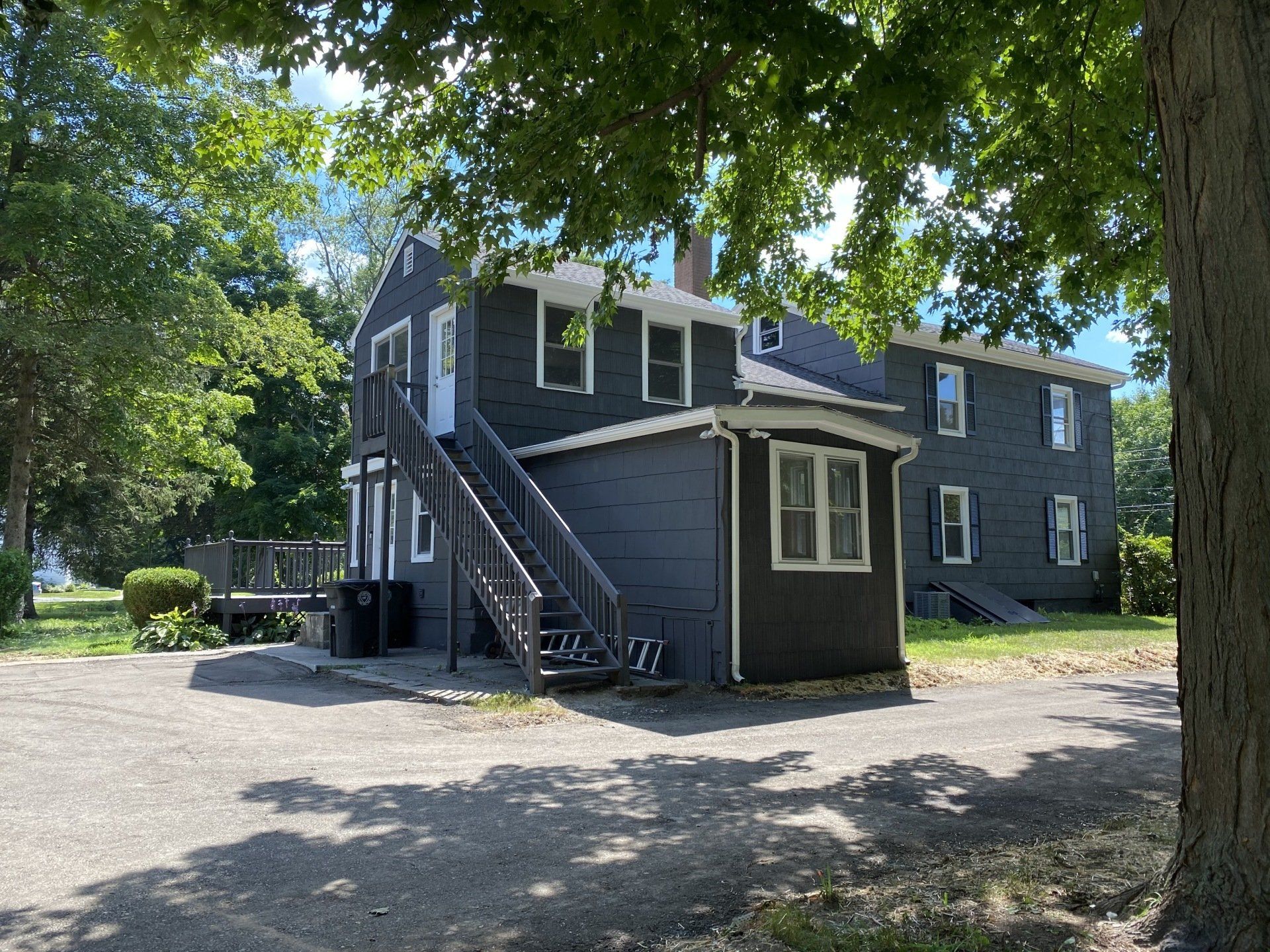 Two-story gray house with exterior stairs, gravel drive, and trees in a sunny setting.