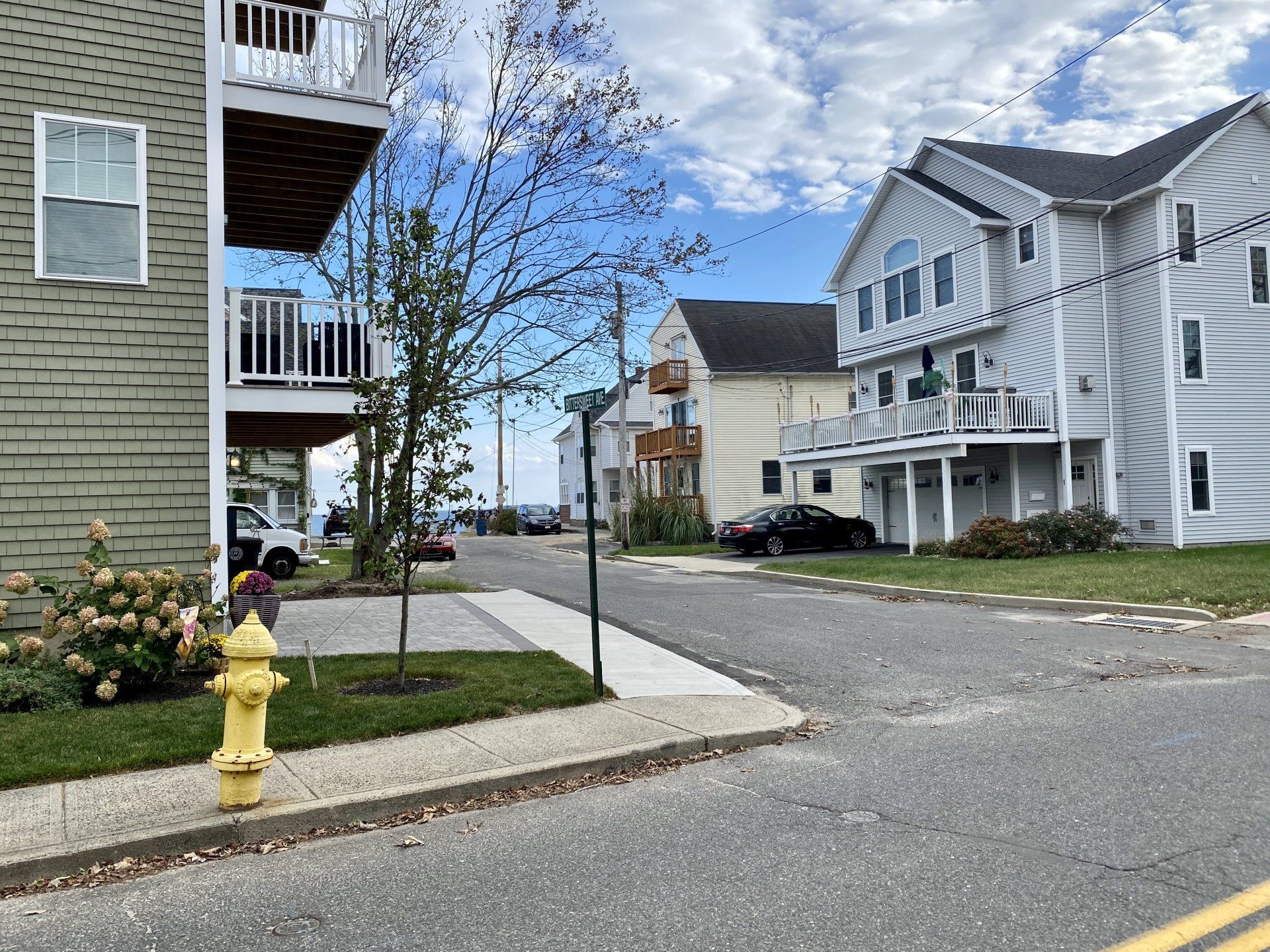 Street view: houses, parked cars, sidewalk, yellow fire hydrant, cloudy sky.