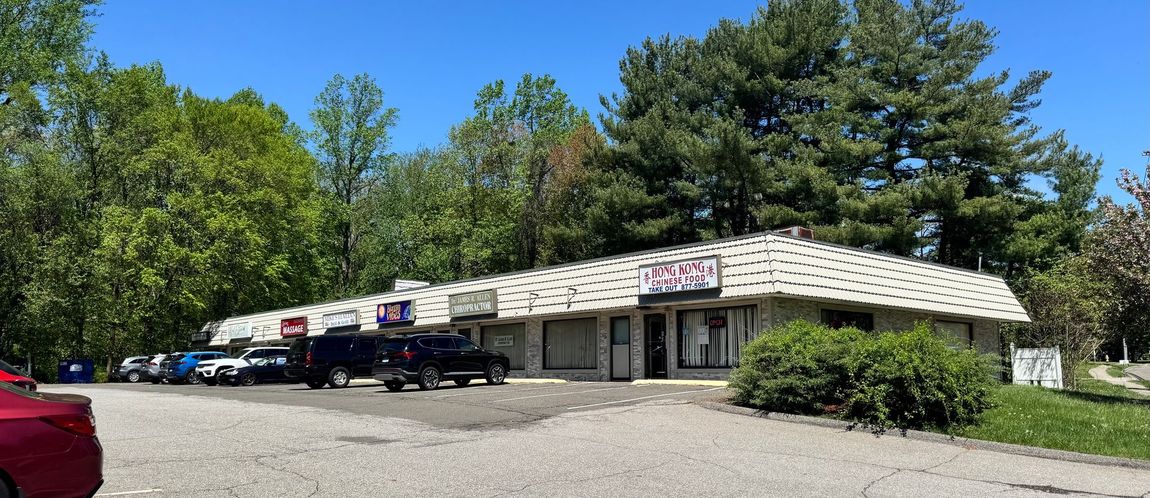 Strip mall with parking area on a sunny day. Trees in the background, blue sky.