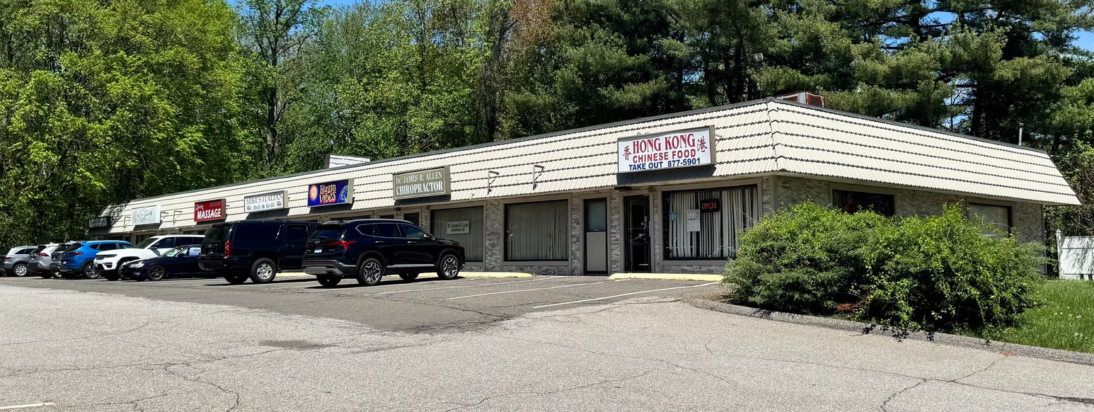 Strip mall with parking area on a sunny day. Trees in the background, blue sky.