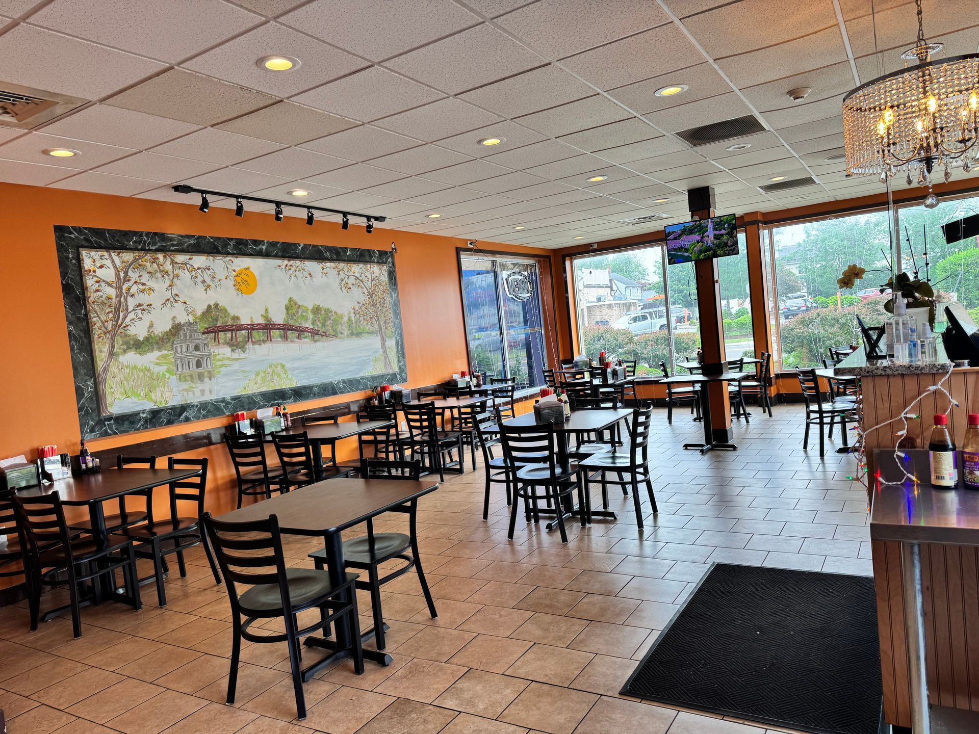 Restaurant interior with tables, chairs, orange wall with artwork, and large windows.