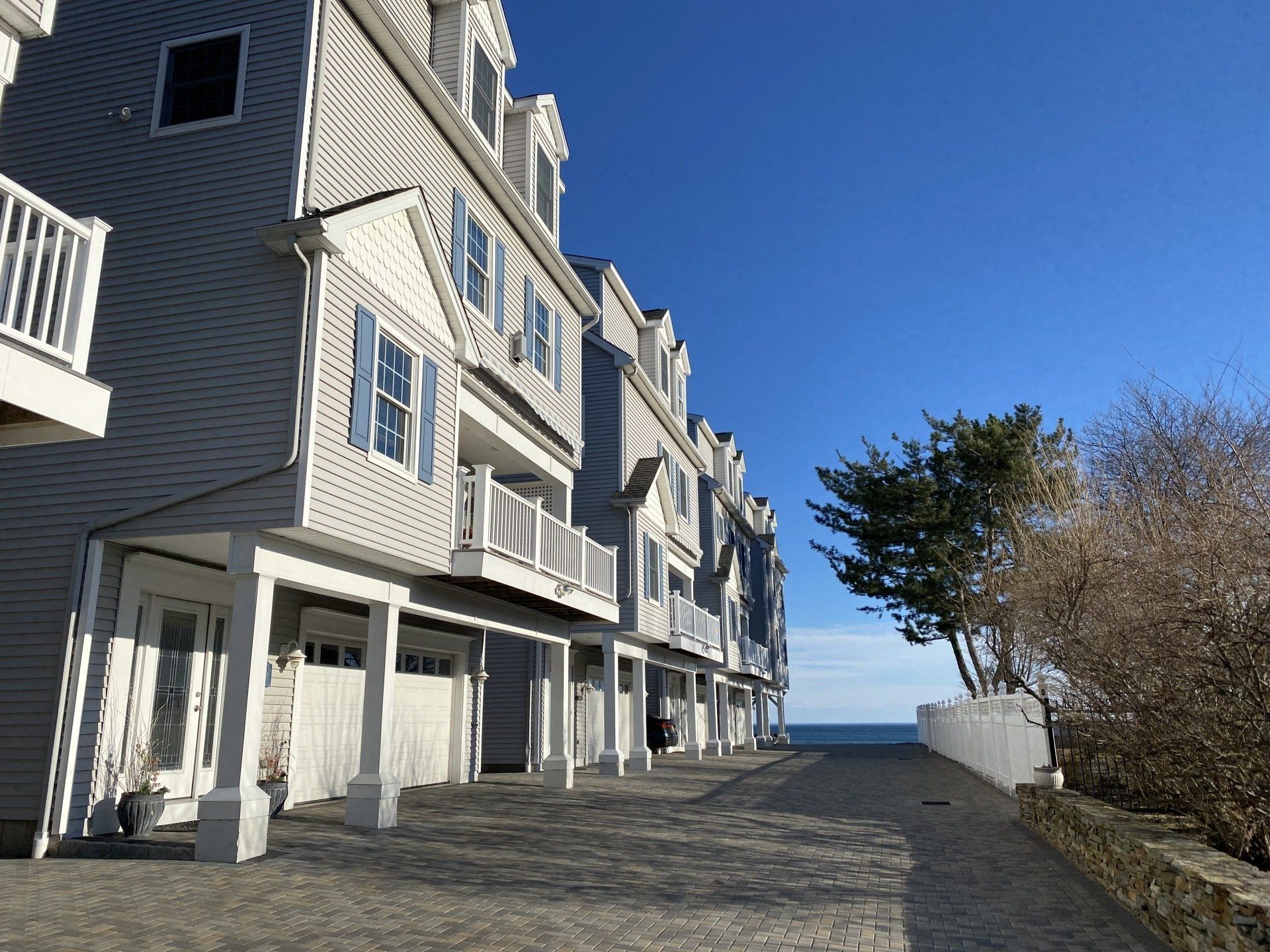 Row of gray townhouses with white garages and balconies, facing the ocean under a blue sky.