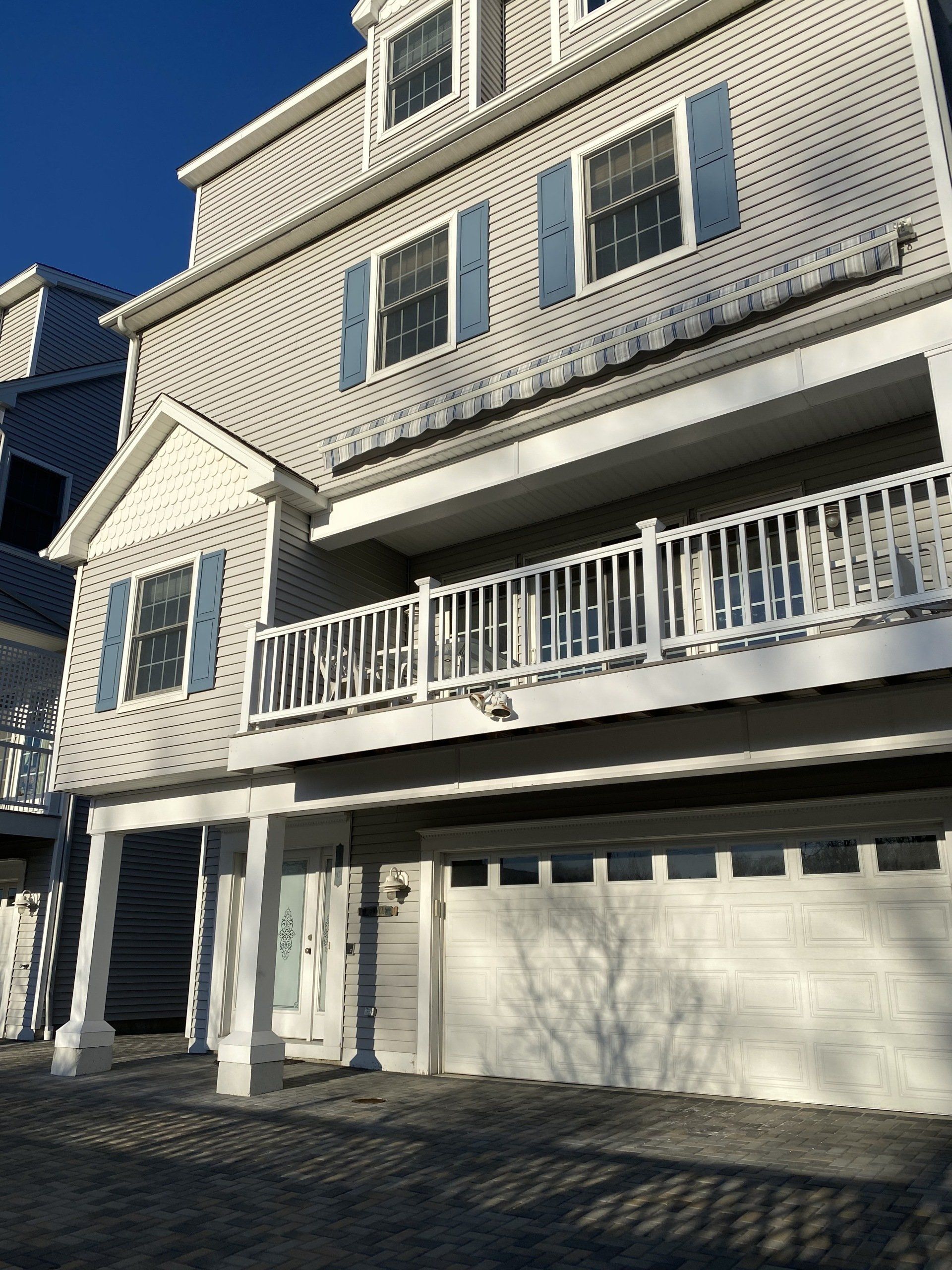 Gray multi-story house with blue shutters, a white balcony, and a garage door, under a blue sky.